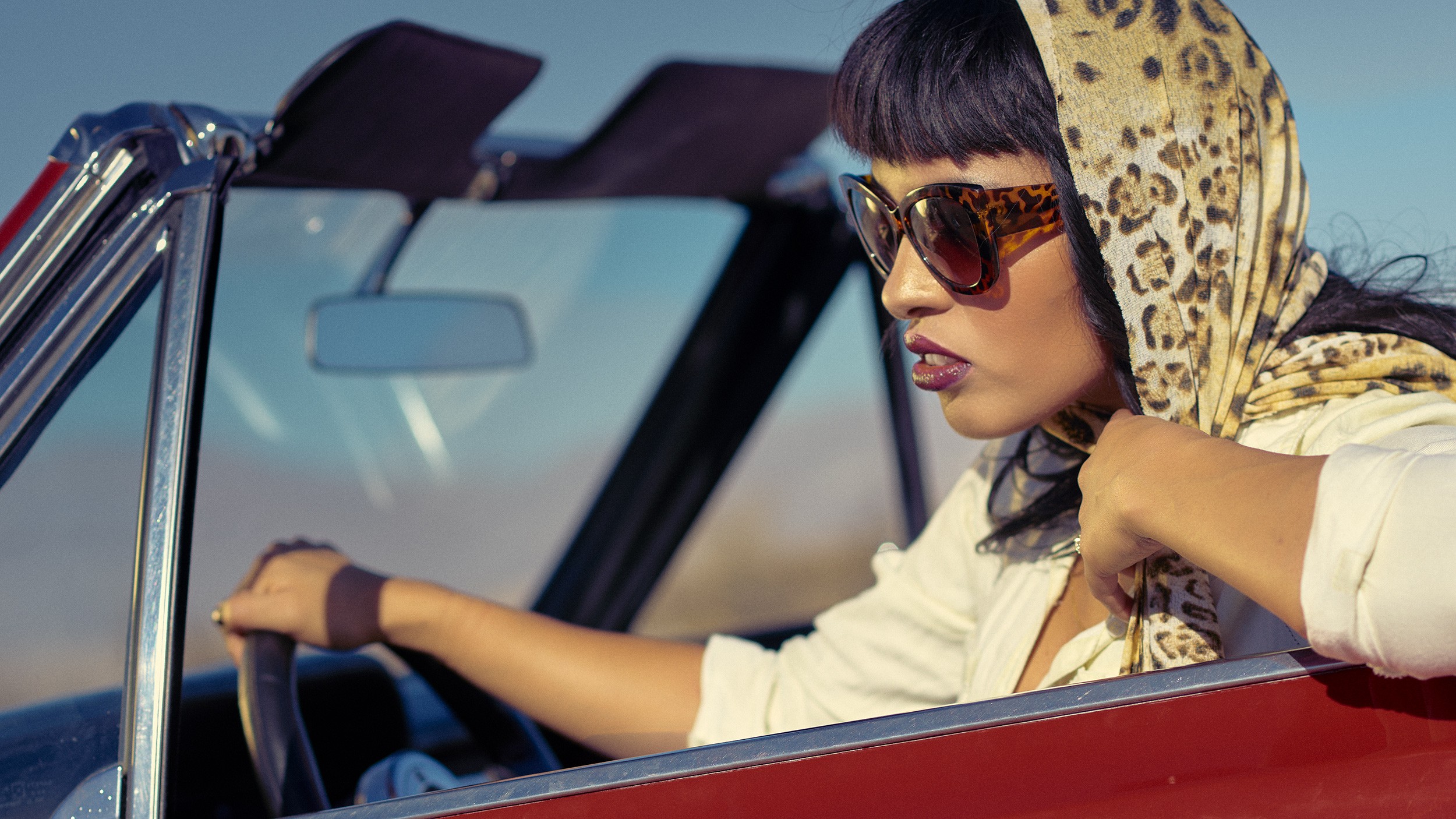 Woman with sunglasses and a leopard print headscarf driving a convertible car, looking to the side.