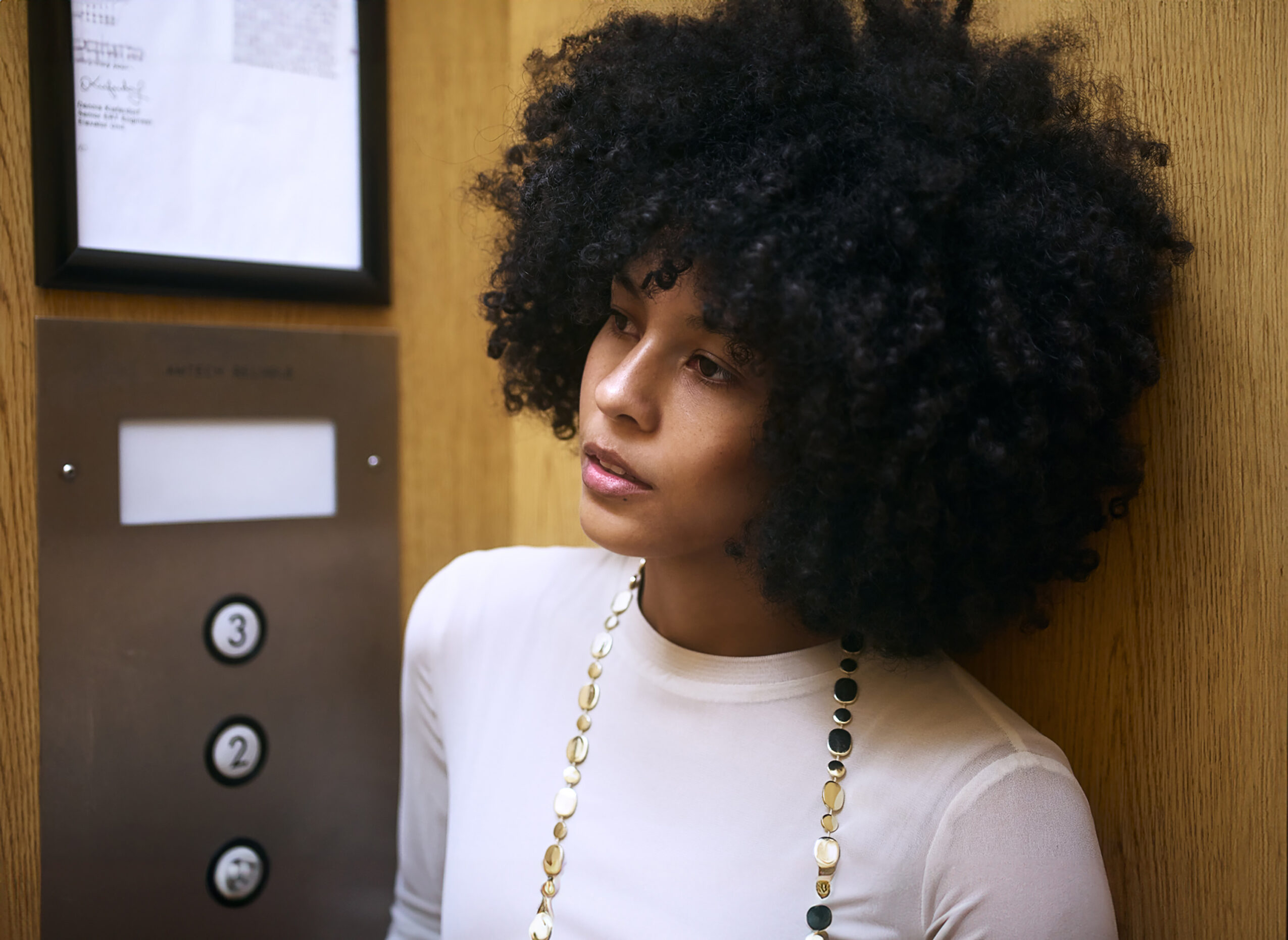 Woman with curly hair leaning against a wooden wall, wearing a white top and a beaded necklace, in an elevator.