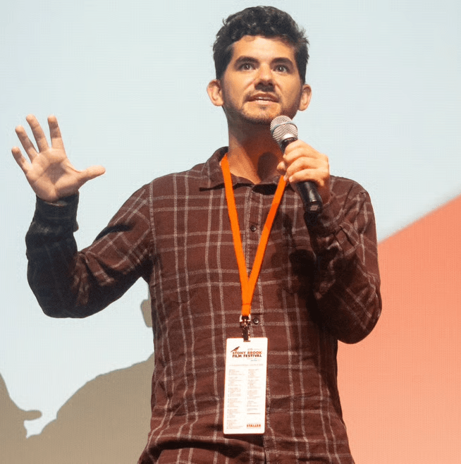 Man speaking into a microphone, gesturing with his left hand, wearing a lanyard and badge, standing against a plain background.