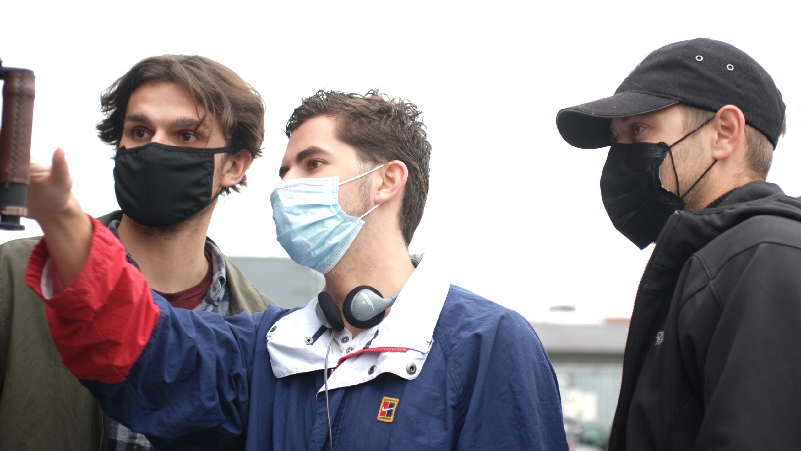 Three people wearing face masks, one with headphones, outdoors, with a cloudy sky background.