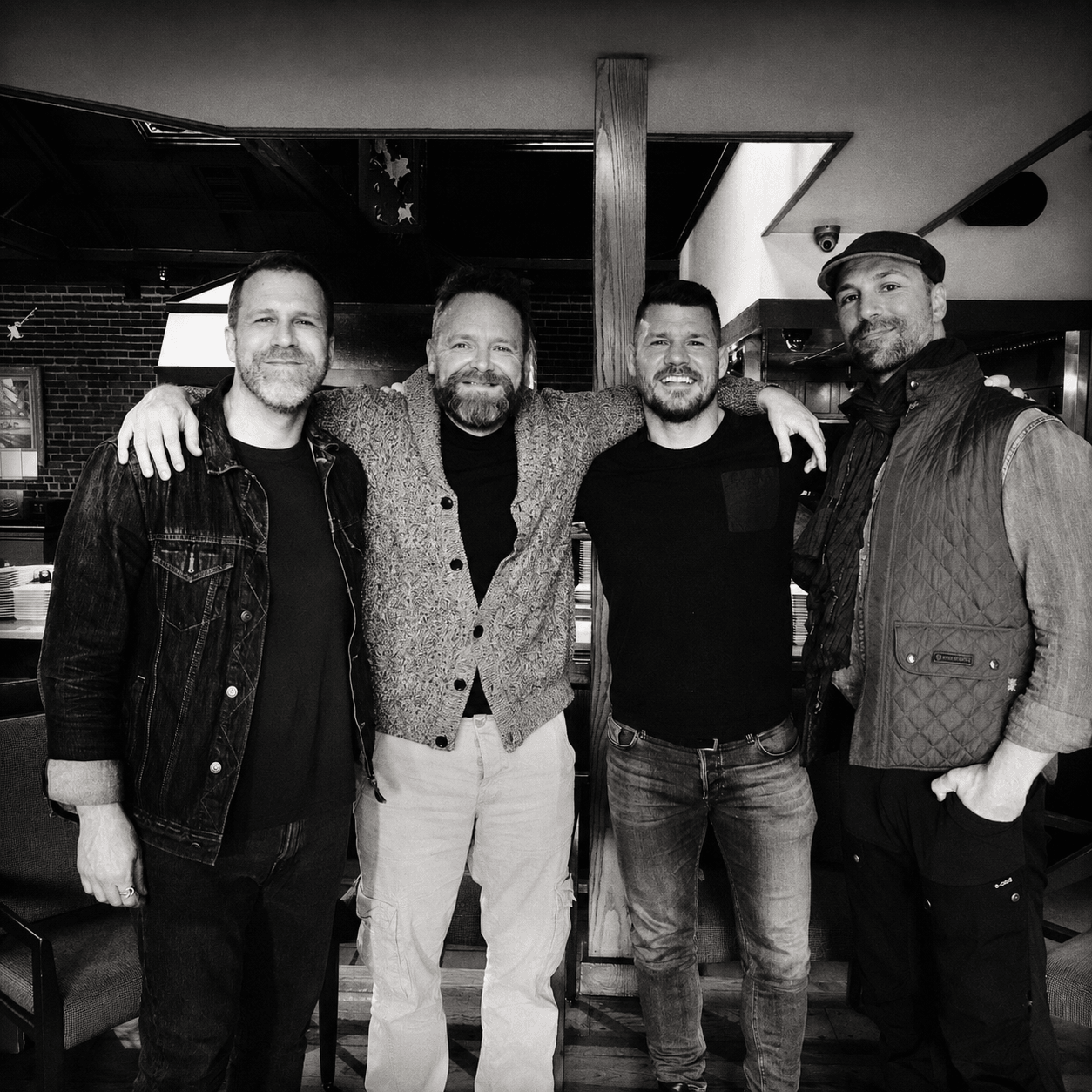 Four men standing together indoors, smiling, with arms around each other, in a casual setting.
