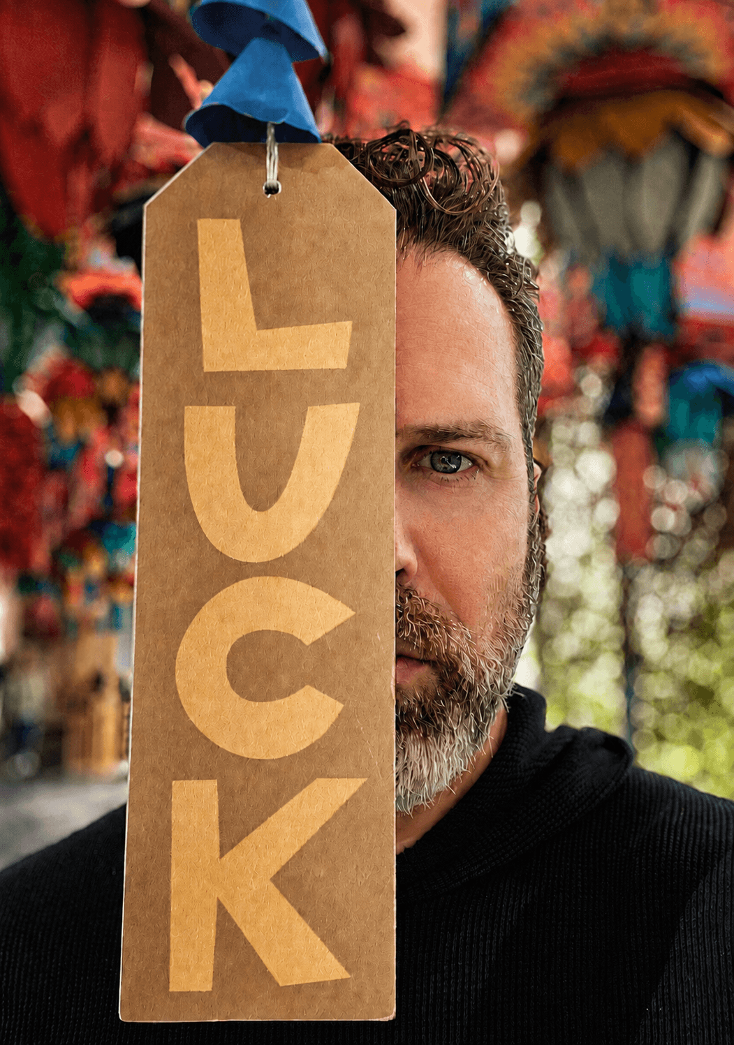 Half of a man's face with a beard and curly hair, with colorful background, and a large brown tag with the word 'LUCK' hanging from above.