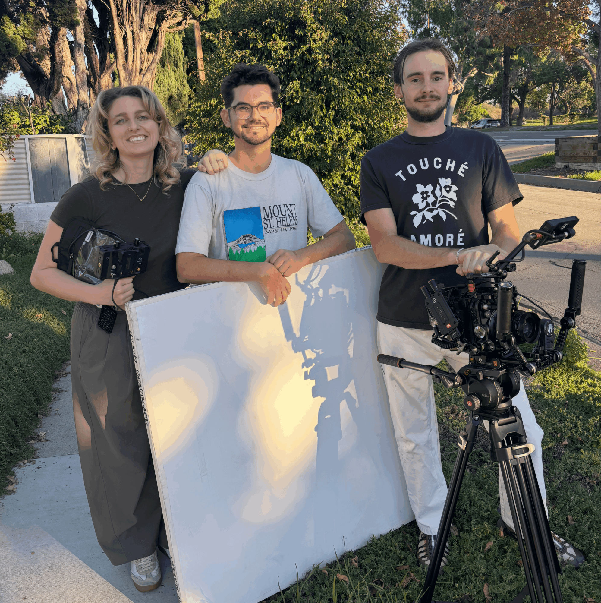 Three people outdoors, two holding cameras, one with a large white board, trees and sunlight in background.