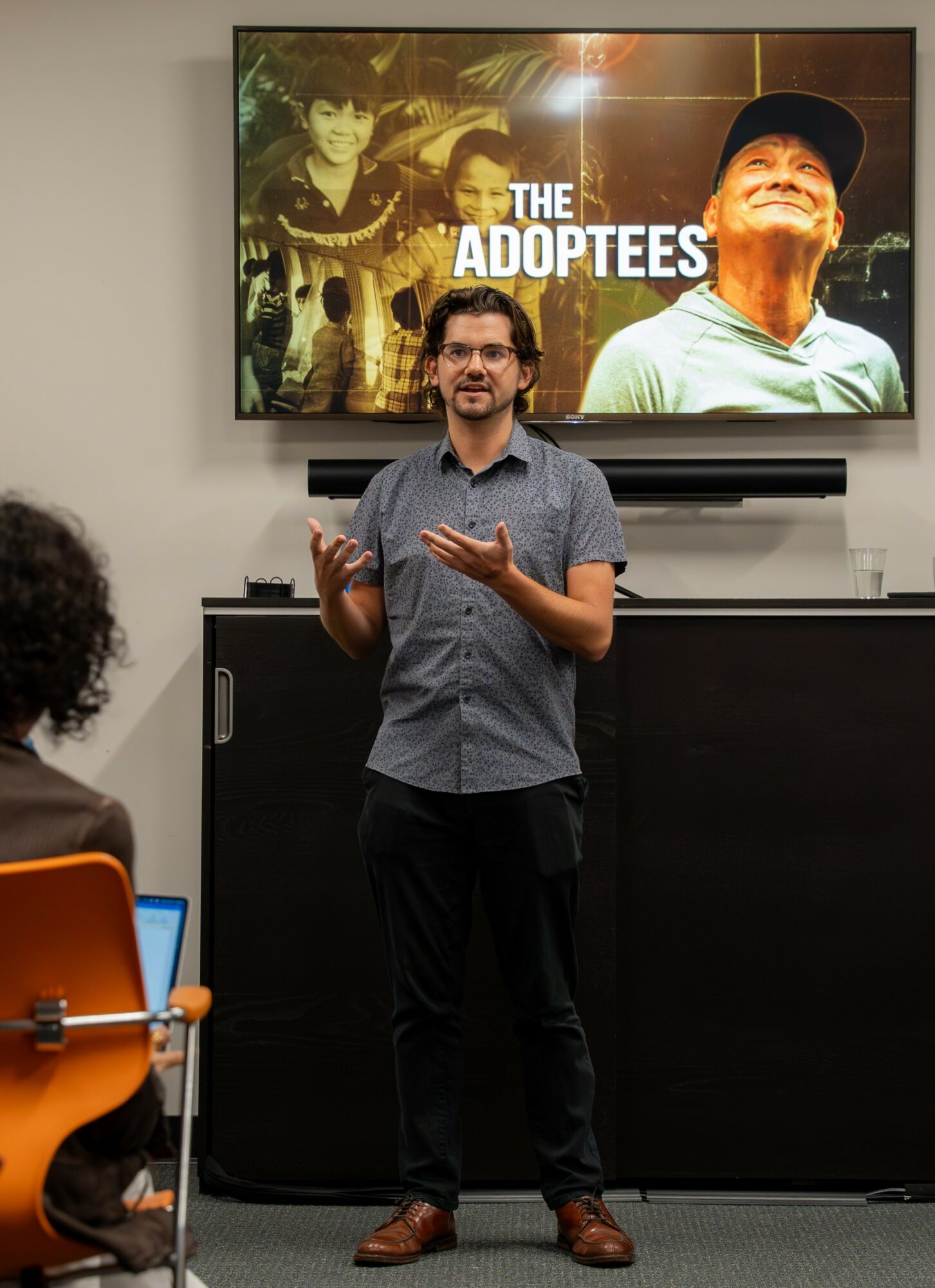 Person standing and speaking in front of a screen displaying the title 'The Adoptees' with a group photo, in a room with an audience.