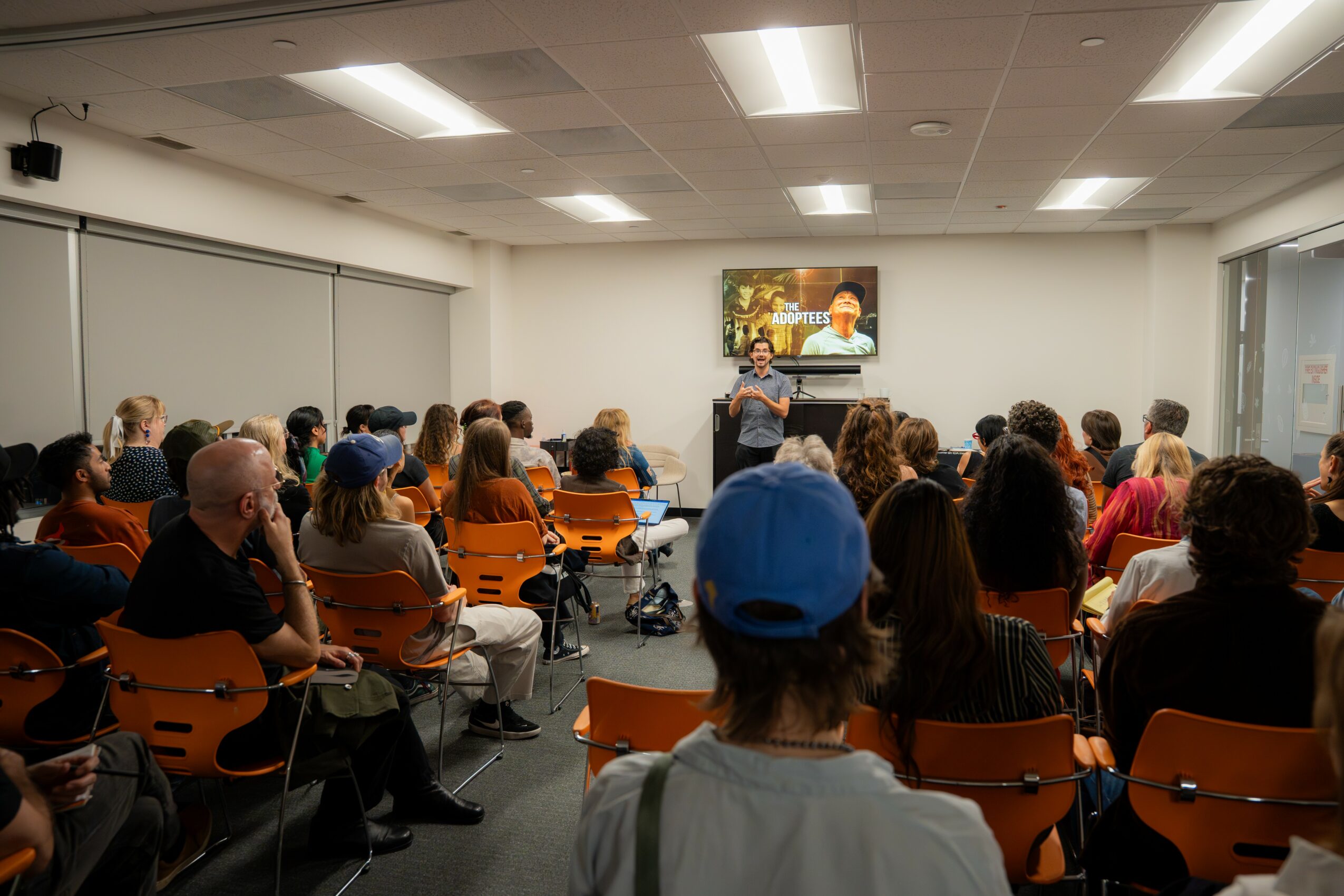 People seated in a classroom watching a presentation on a screen, with a teacher at the front.