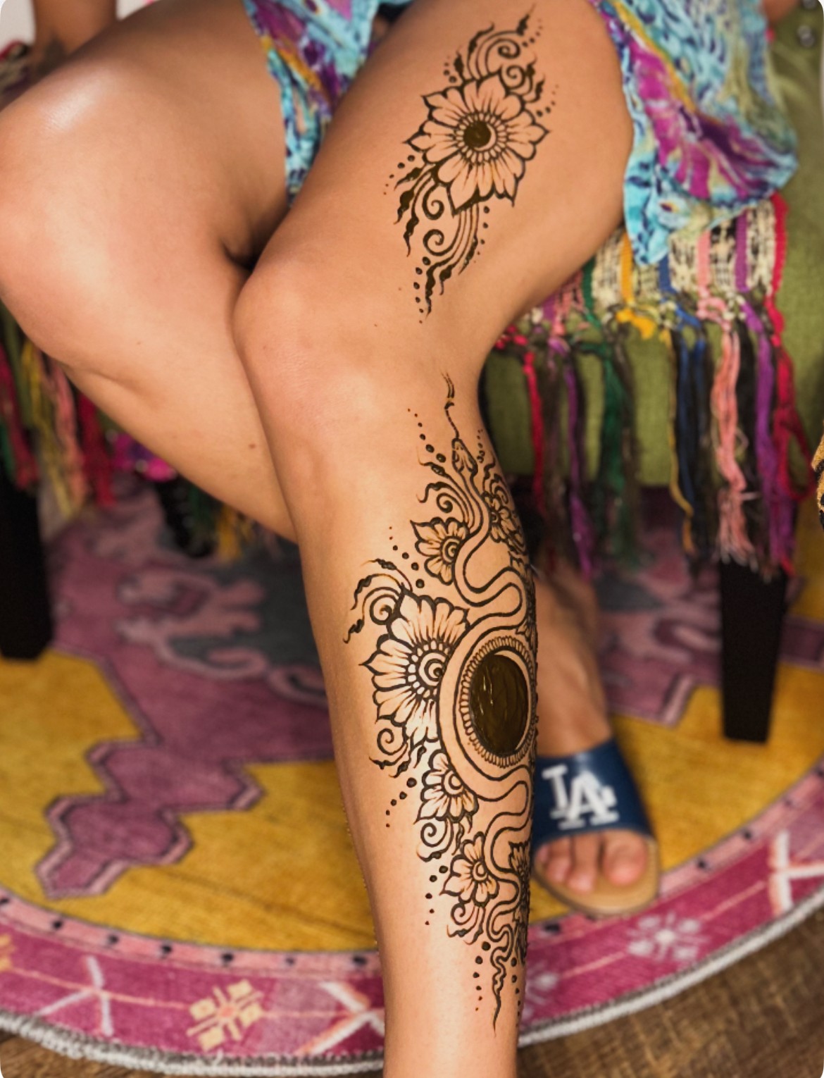 Person with a large floral and circular tattoo on their leg, sitting on a colorful rug with a multicolored fabric background.