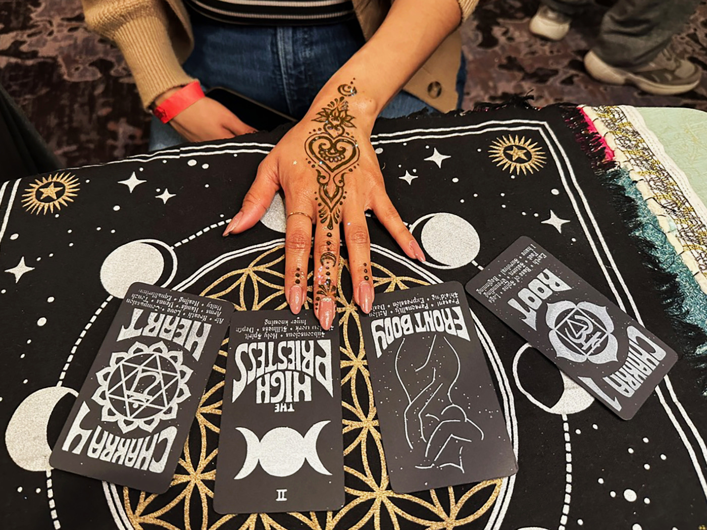 Person's hand with henna tattoo resting on a black cloth with celestial designs, tarot cards spread out, and a fringed edge.
