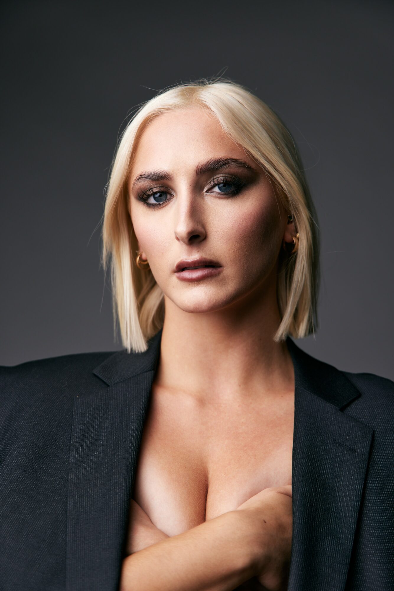 Portrait of a woman with platinum blonde hair, wearing a black blazer, looking directly at the camera against a dark background.
