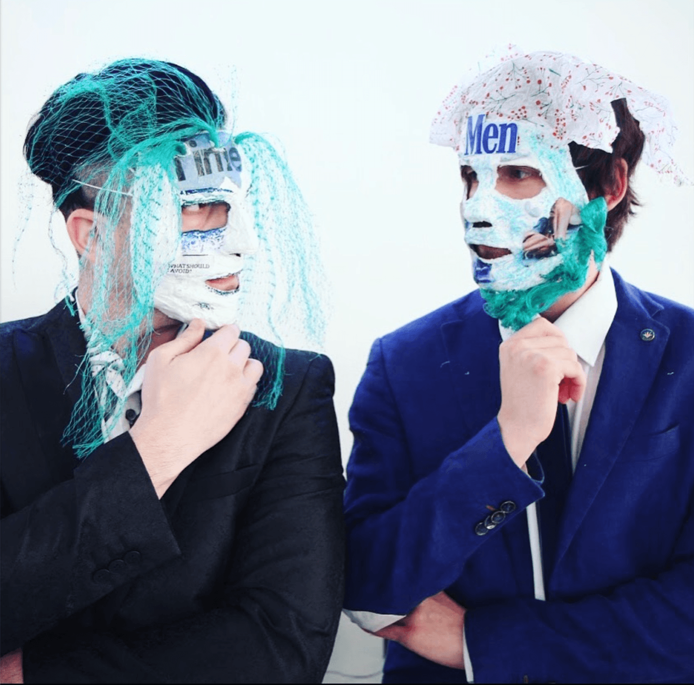 Two people wearing masks and costumes, one with teal hair and the other with a newspaper hat, facing each other.