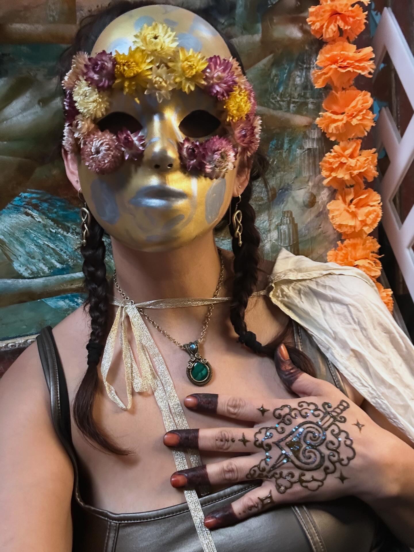 Person wearing a gold mask with floral decorations, braided hair, jewelry, and a henna design on hand, with orange flowers in background.
