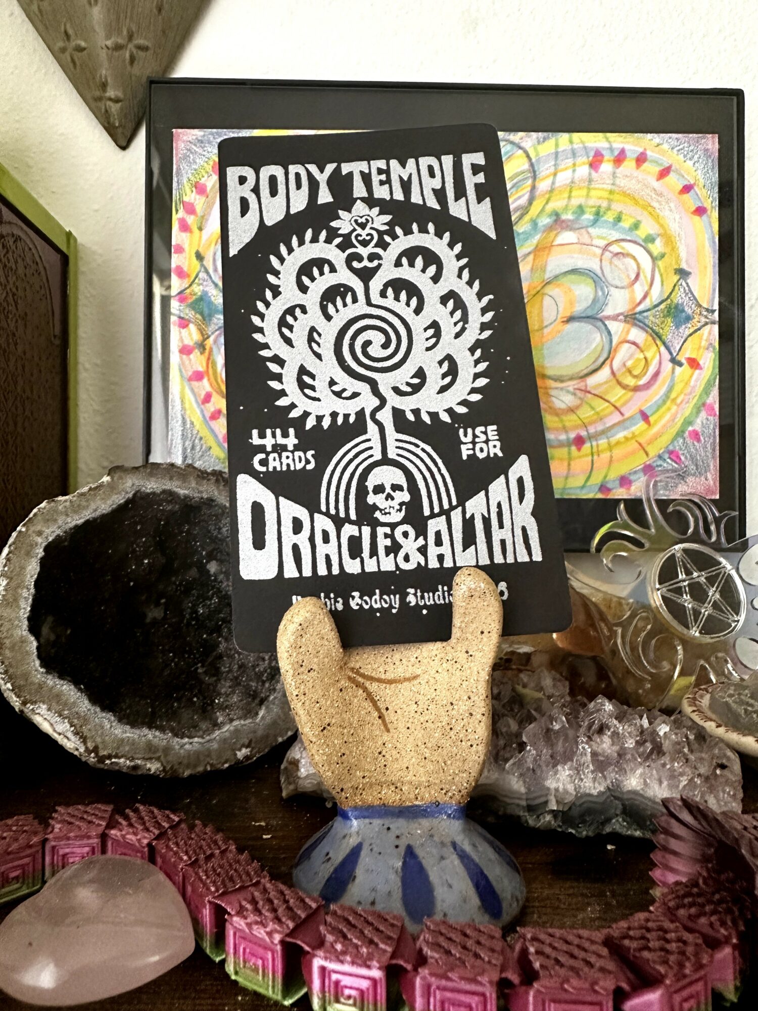 Decorative display with a black and white card, a small ceramic hand, crystals, and colorful artwork in background.