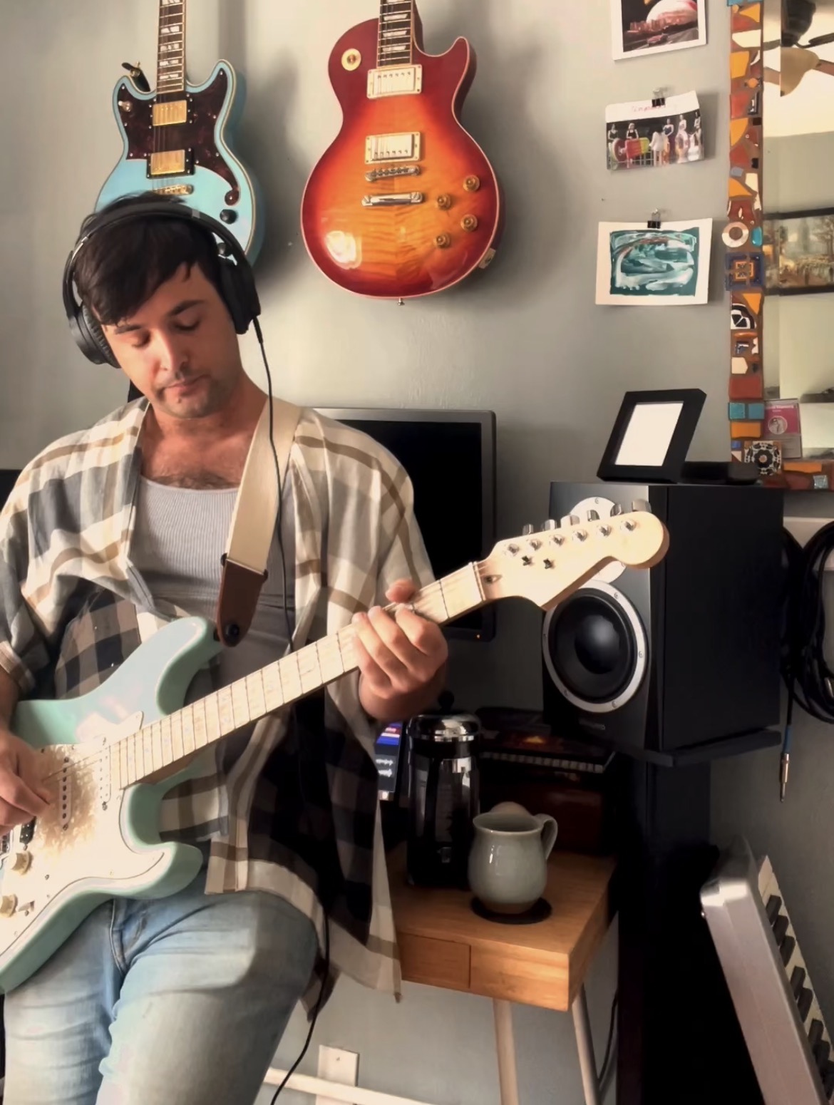 Person playing electric guitar with headphones in a room decorated with guitars and photos on the wall.