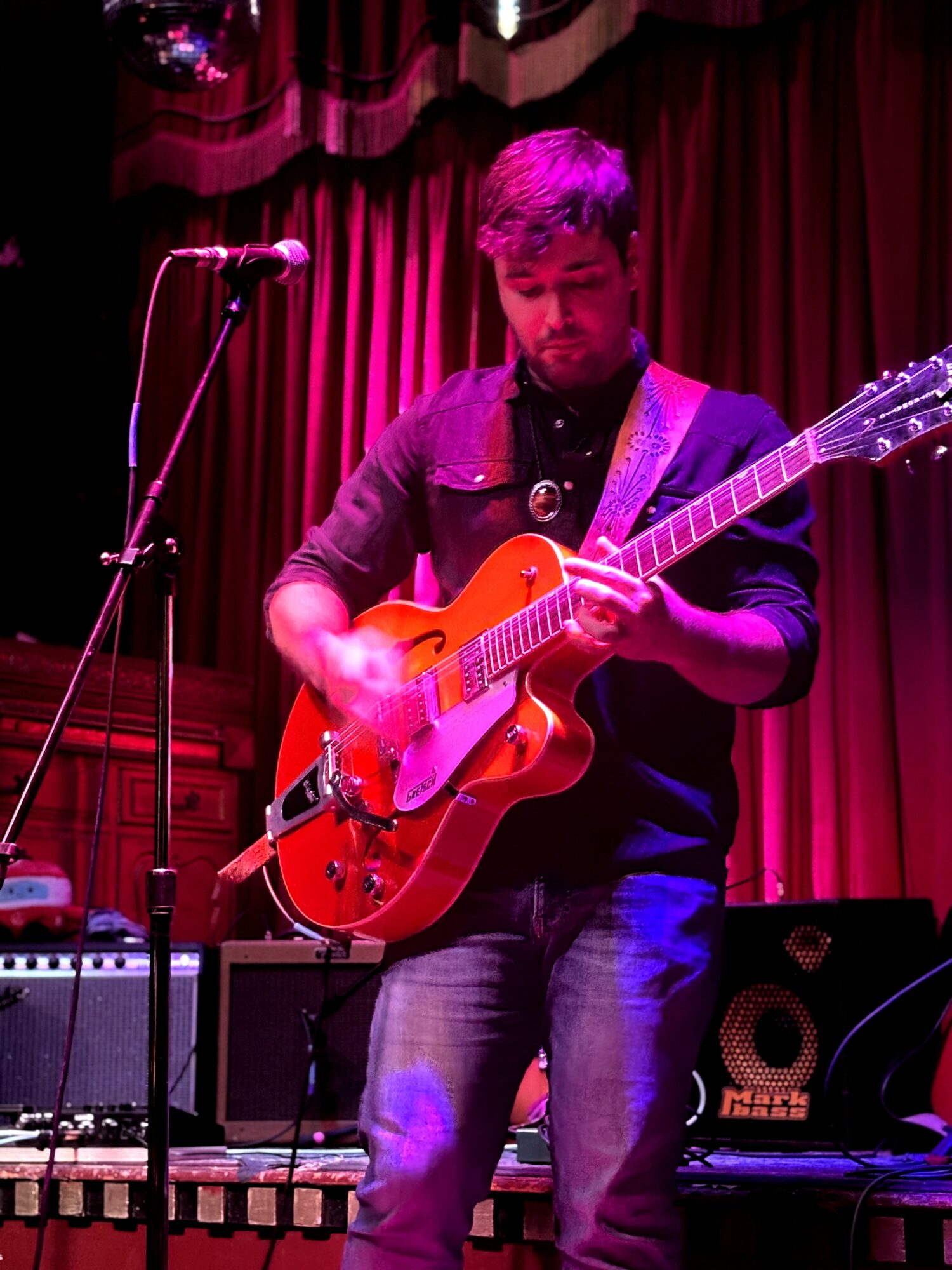 Man playing an orange electric guitar on stage with red curtains behind him.