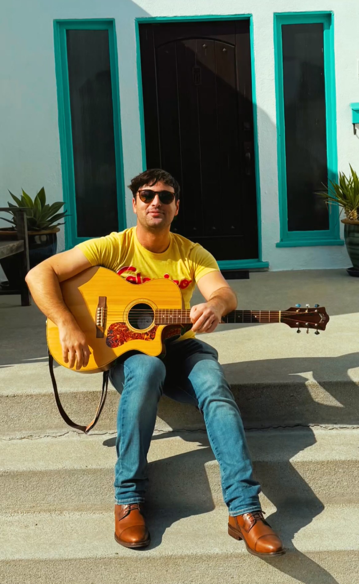 Person sitting on steps playing guitar in front of a building with teal window frames and plants on either side.