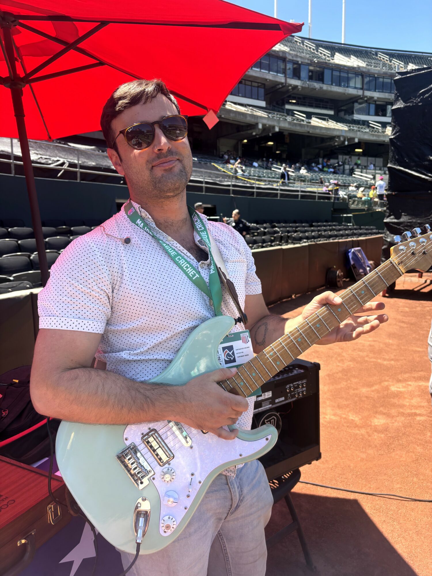 Man with sunglasses holding a white electric guitar under a red umbrella at a marina with boats and buildings in the background.