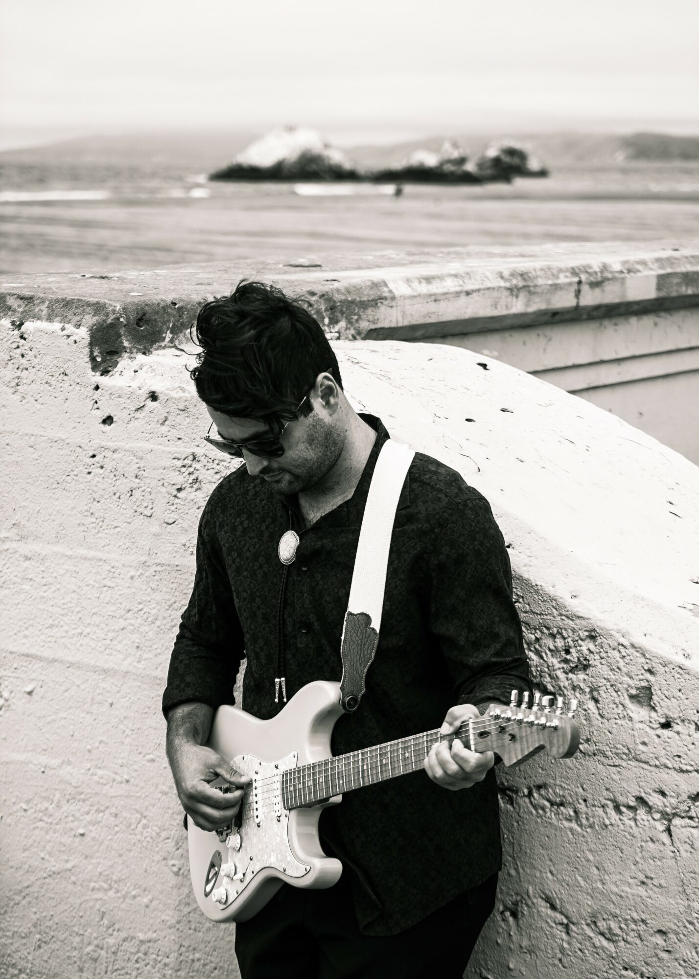 Person with dark hair and sunglasses playing an electric guitar against a white wall, with a ship in the background.