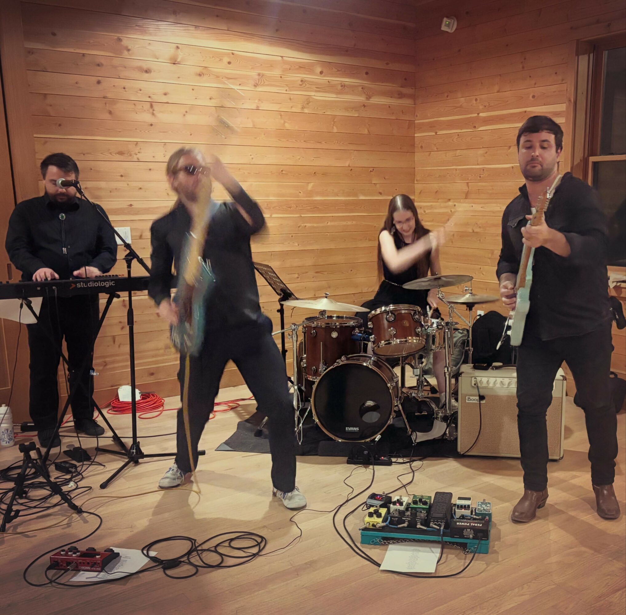 Four musicians perform in a wooden room, with two playing guitars, one on drums, and one on keyboard, all engaged in music.