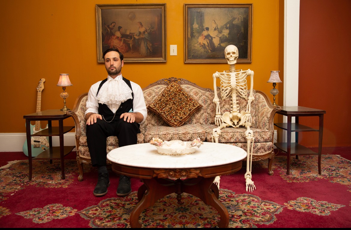Man sitting on a sofa with a skeleton, in a room with paintings, lamps, and a patterned rug.
