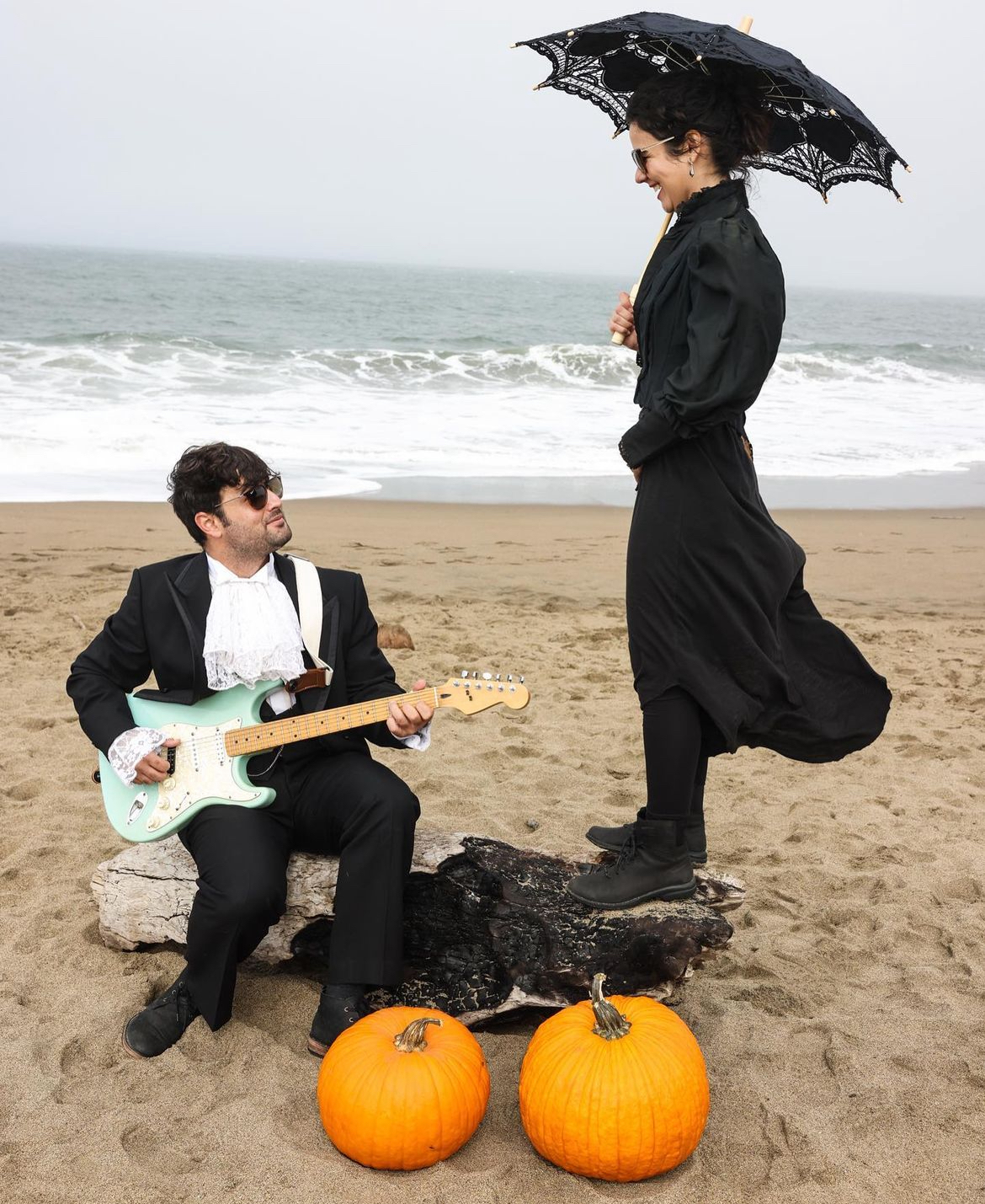 Two people on a beach with pumpkins, one sitting with a guitar and the other standing with an umbrella, overcast sky.