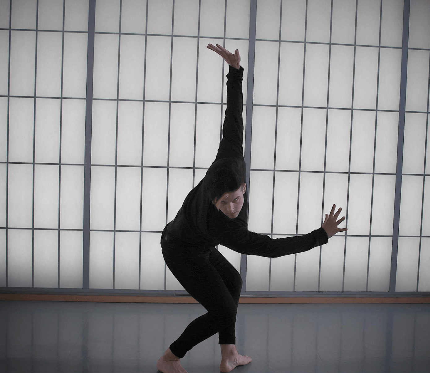 Person in black clothing leaning forward with one arm raised and the other extended sideways, against a grid-patterned wall.
