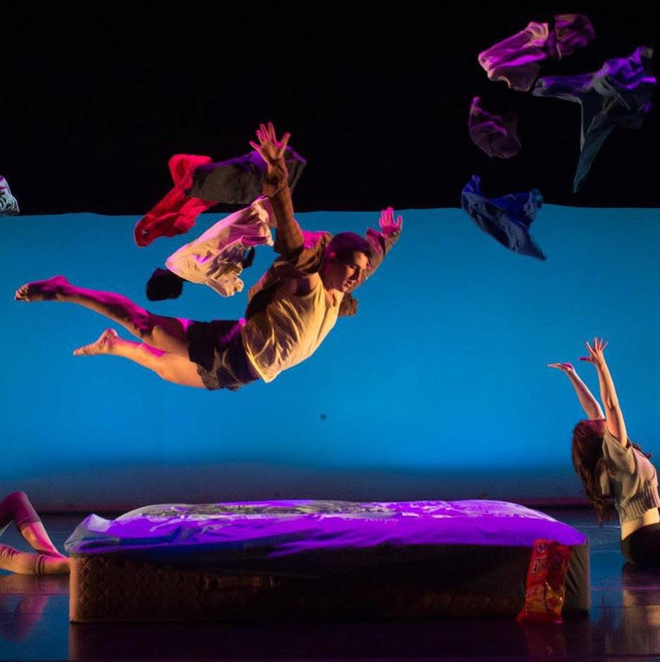 Performer mid-air during a dance routine, with others on stage and colorful costumes, against a blue background.