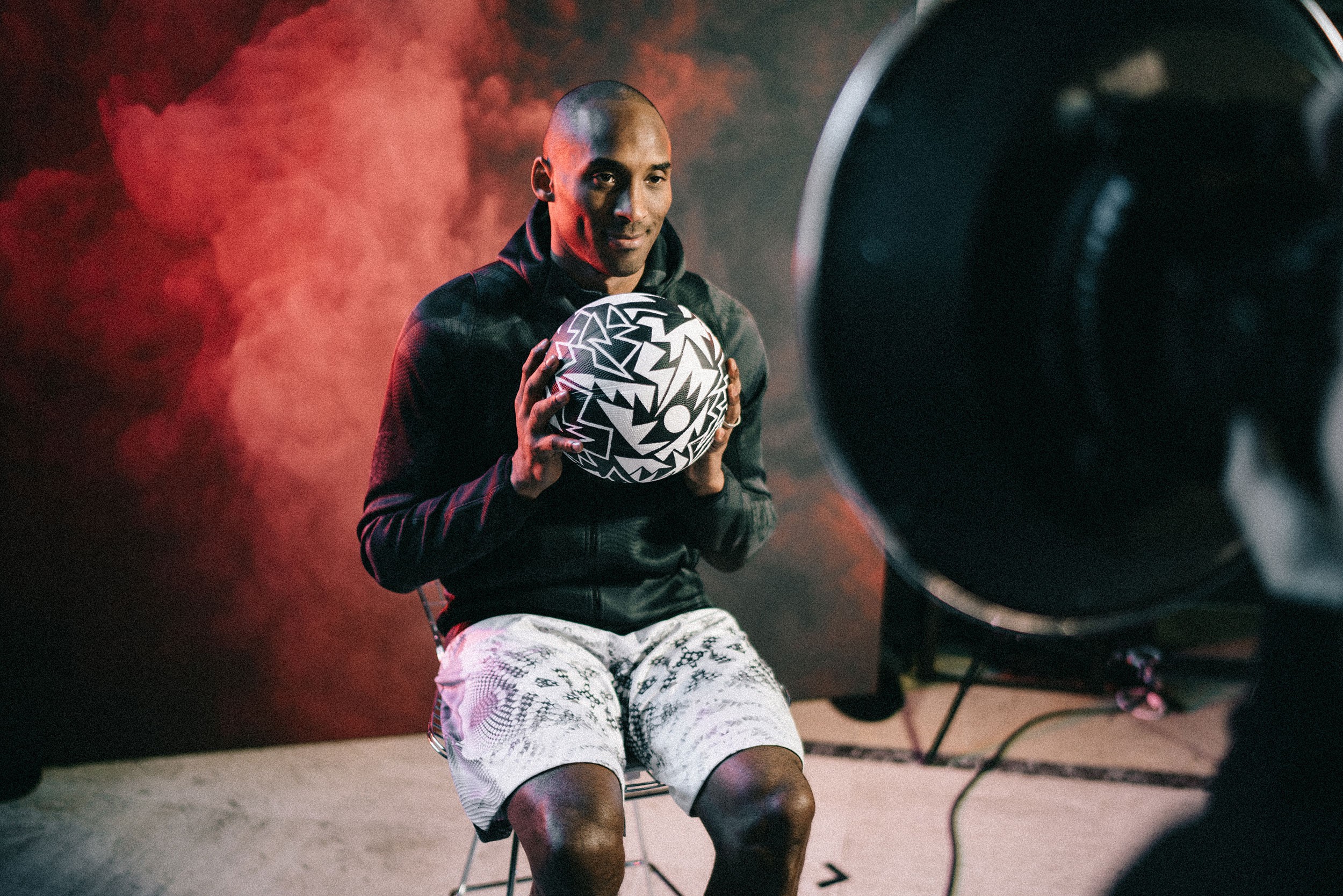 Man sitting on a surface holding a patterned soccer ball, with a camera and studio lighting setup, red smoke in background.