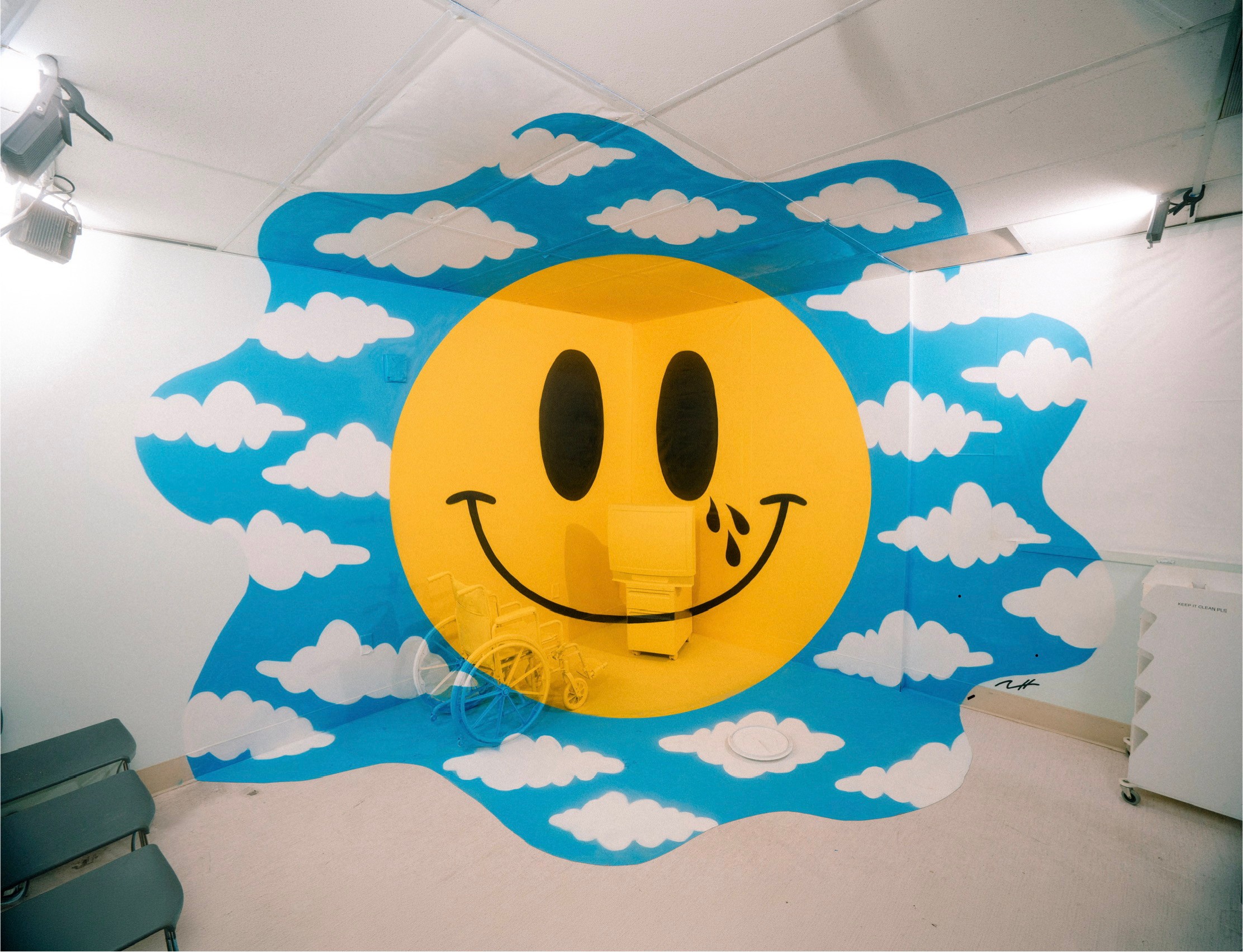 Wall mural features a large yellow smiley face with black eyes and a wide smile, surrounded by blue sky and white clouds.