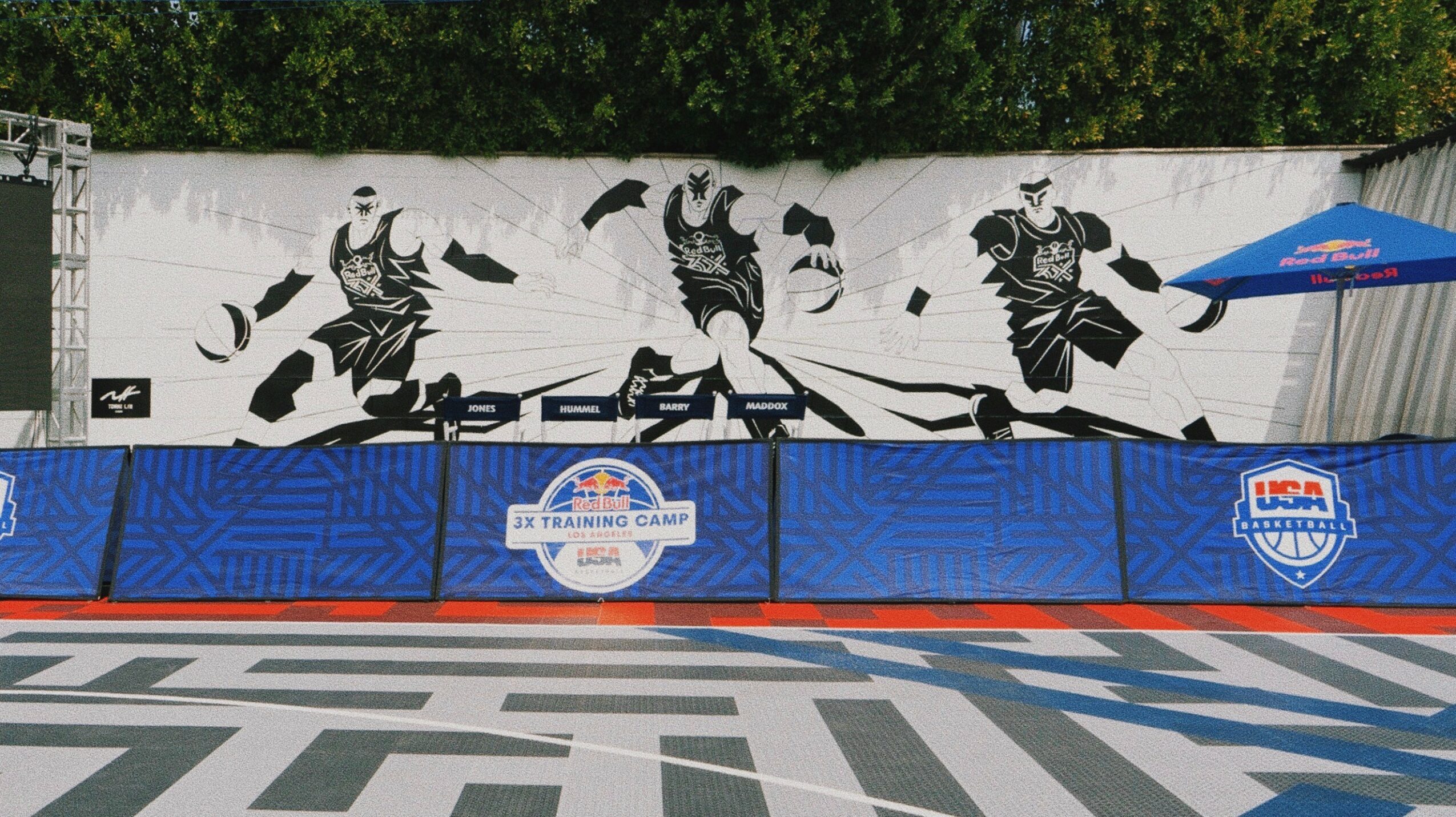 Black and white mural of three dancers on a wall, with a blue barrier and umbrella in front, outdoor setting.