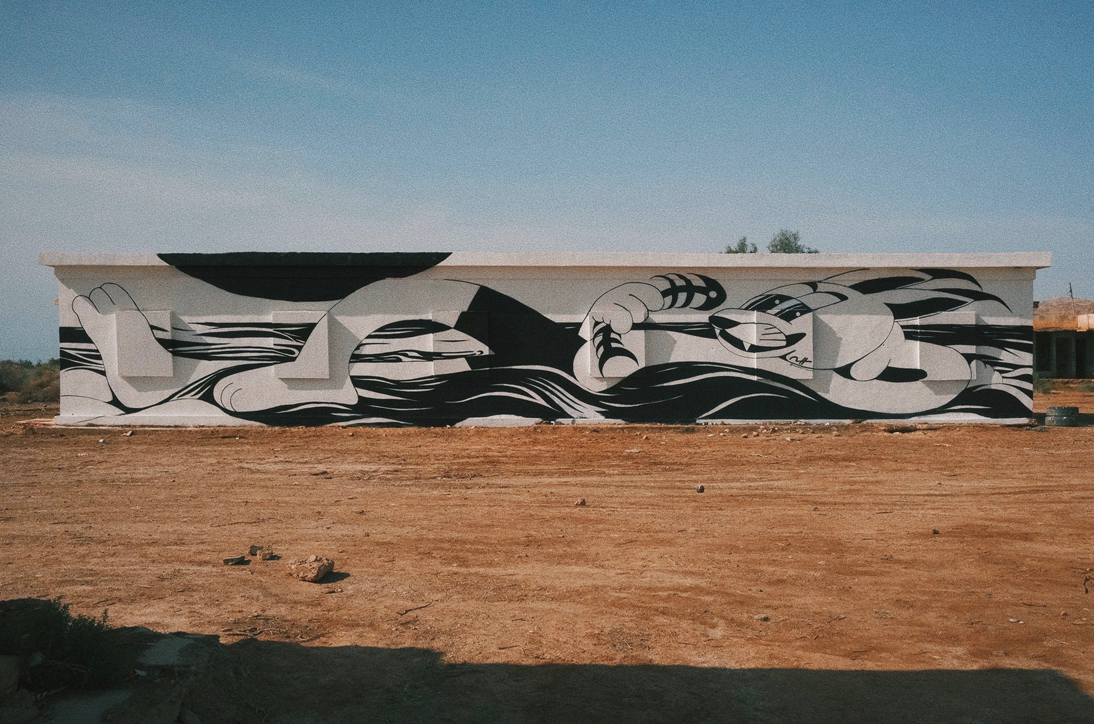 Long building with black and white abstract mural, clear sky, and dirt foreground.