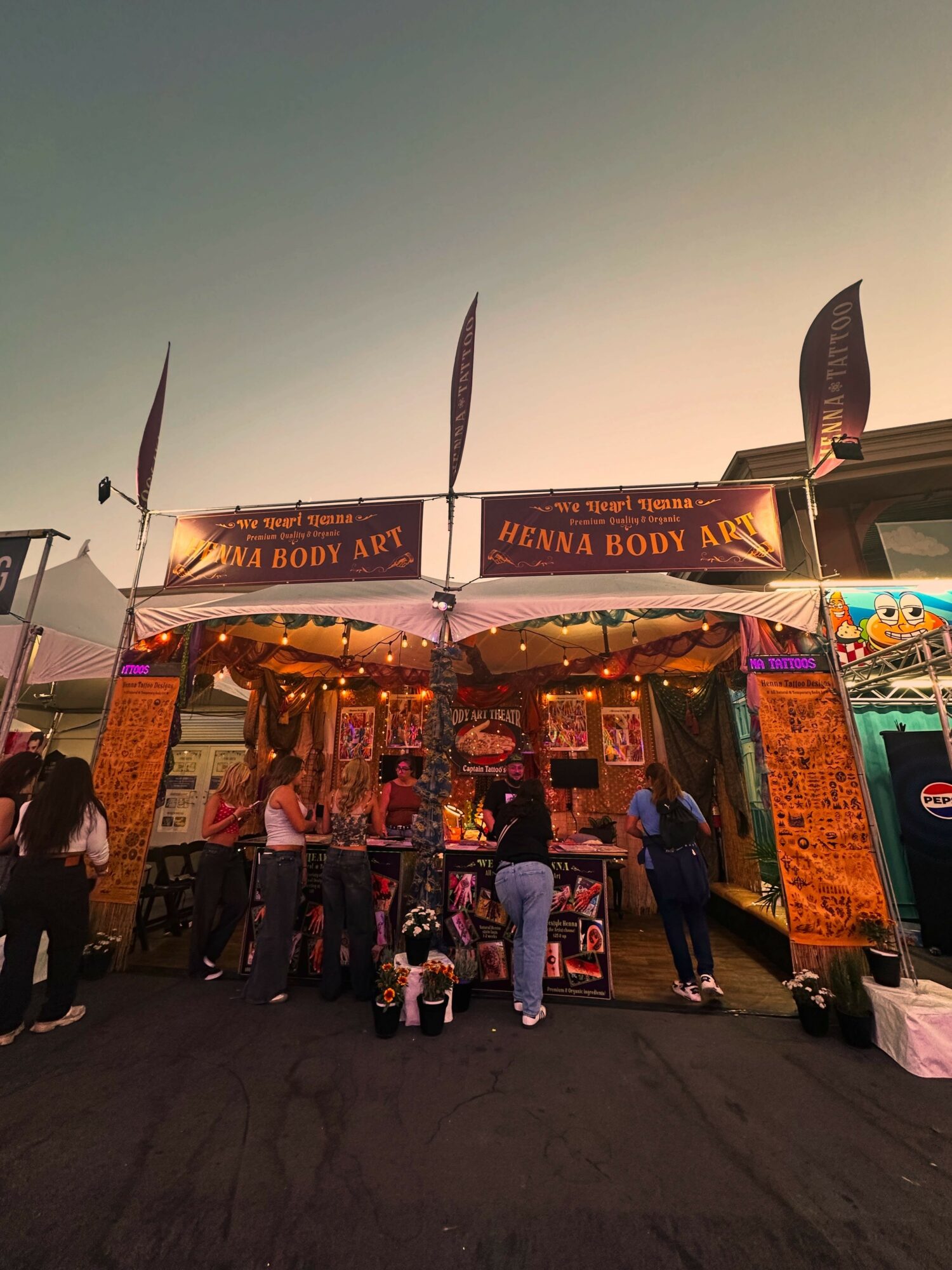 People gather around a booth with a sign reading 'HINNA BODY ART' at an outdoor event during sunset.