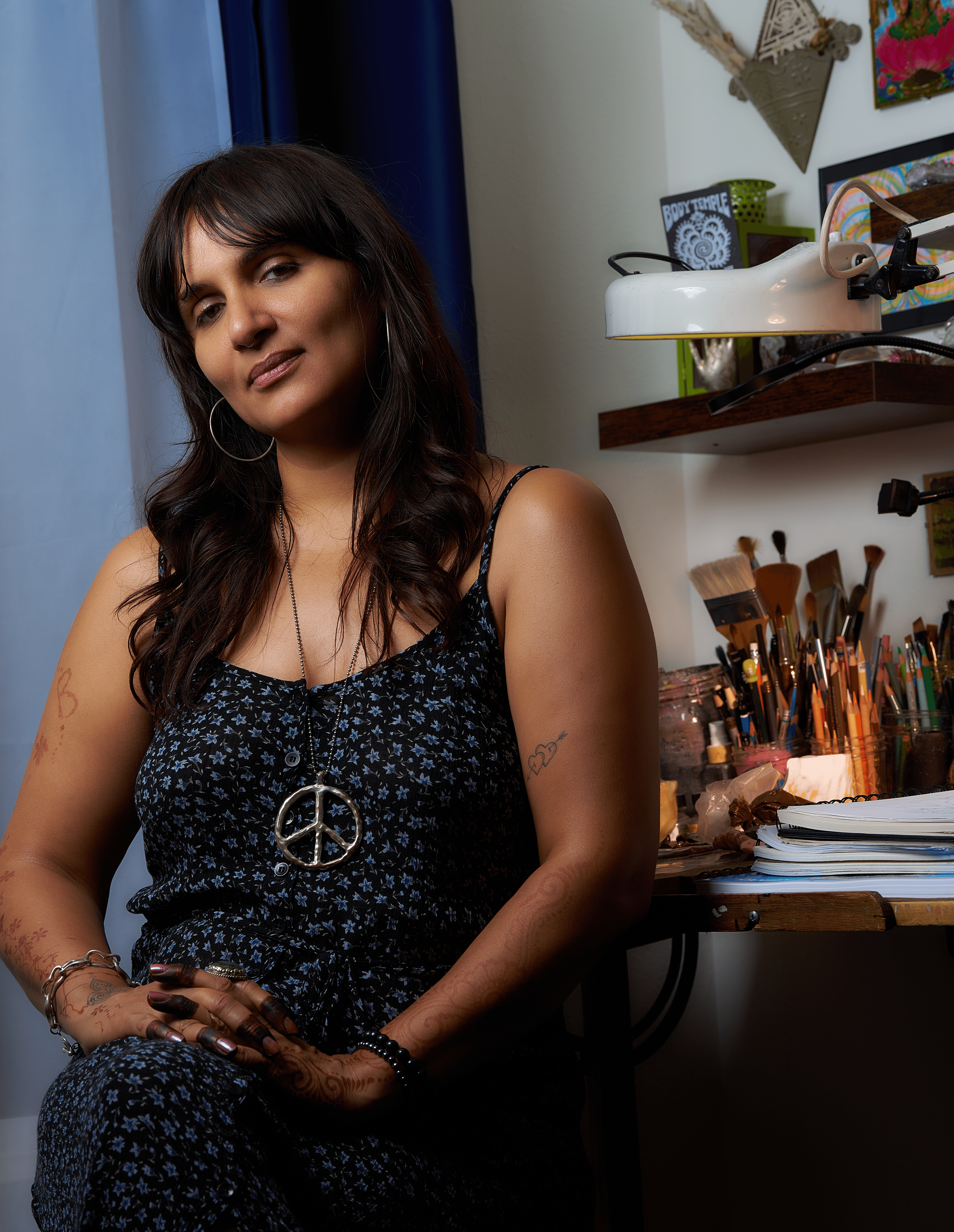 Woman with dark hair and hoop earrings sitting in a room with art supplies and shelves behind her.