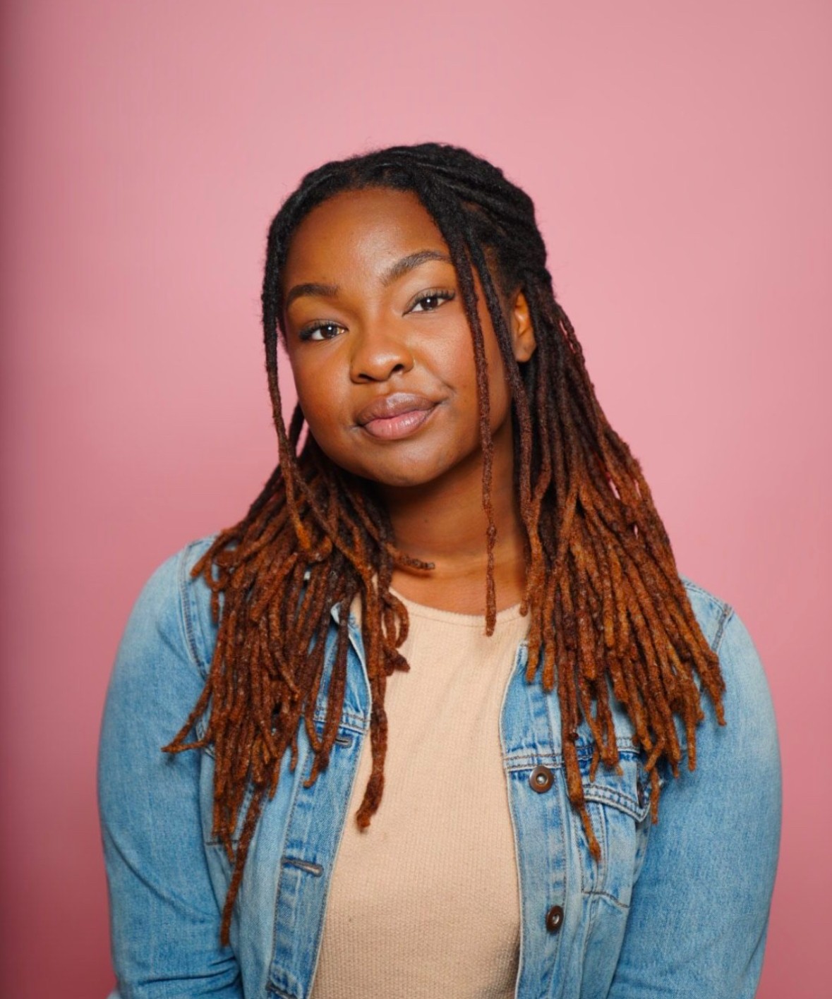 Young woman with long dreadlocks wearing a denim jacket and beige top, standing against pink background.