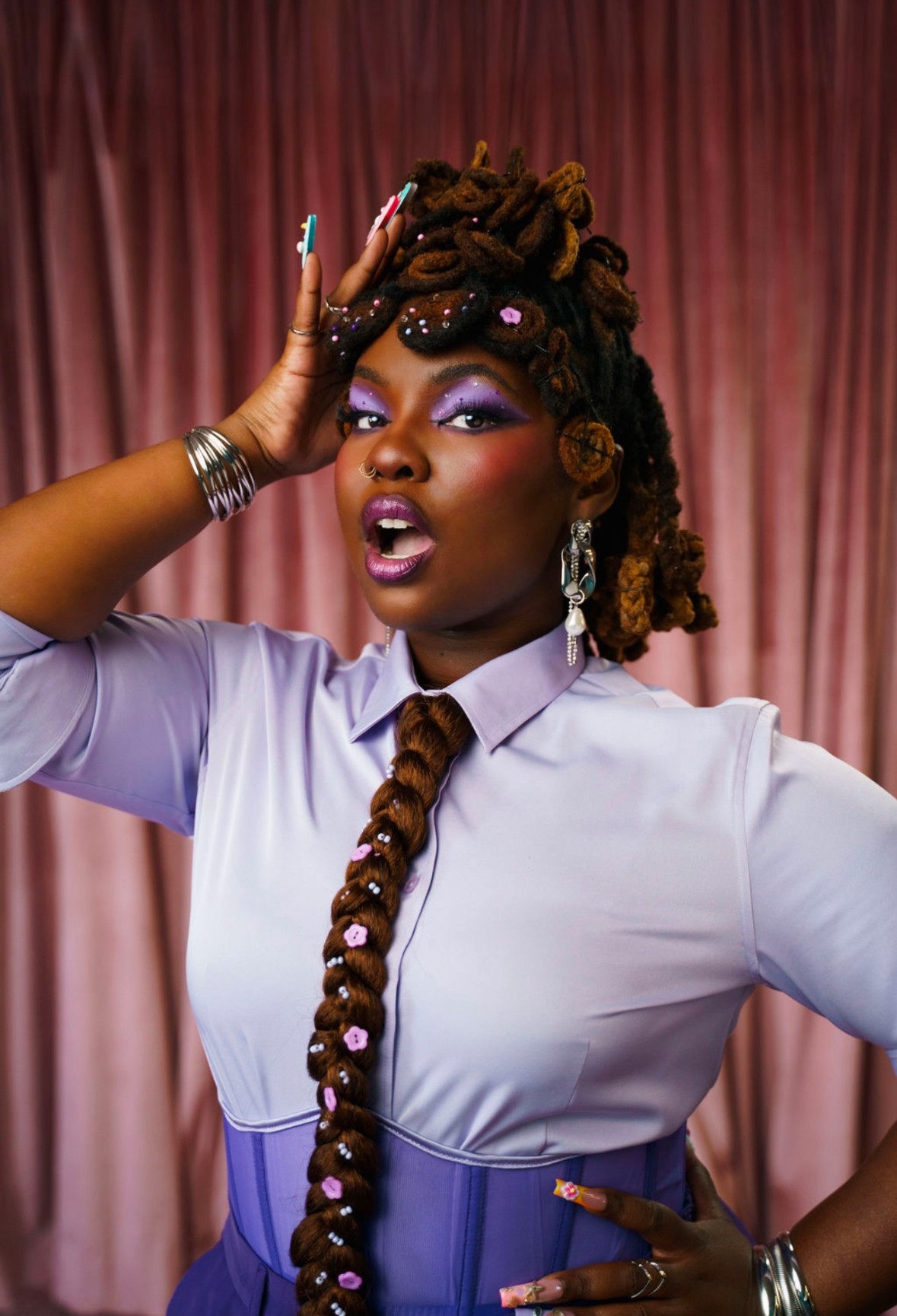 A woman with styled dreadlocks and jewelry poses with her hand on her head against a pink curtain background.