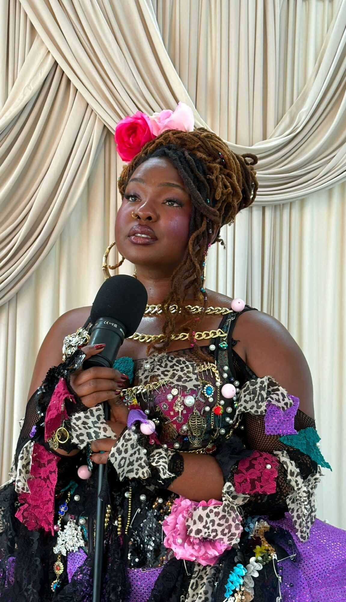 Woman with braided hair wearing colorful accessories and a floral outfit, holding a microphone, standing in front of draped curtains.