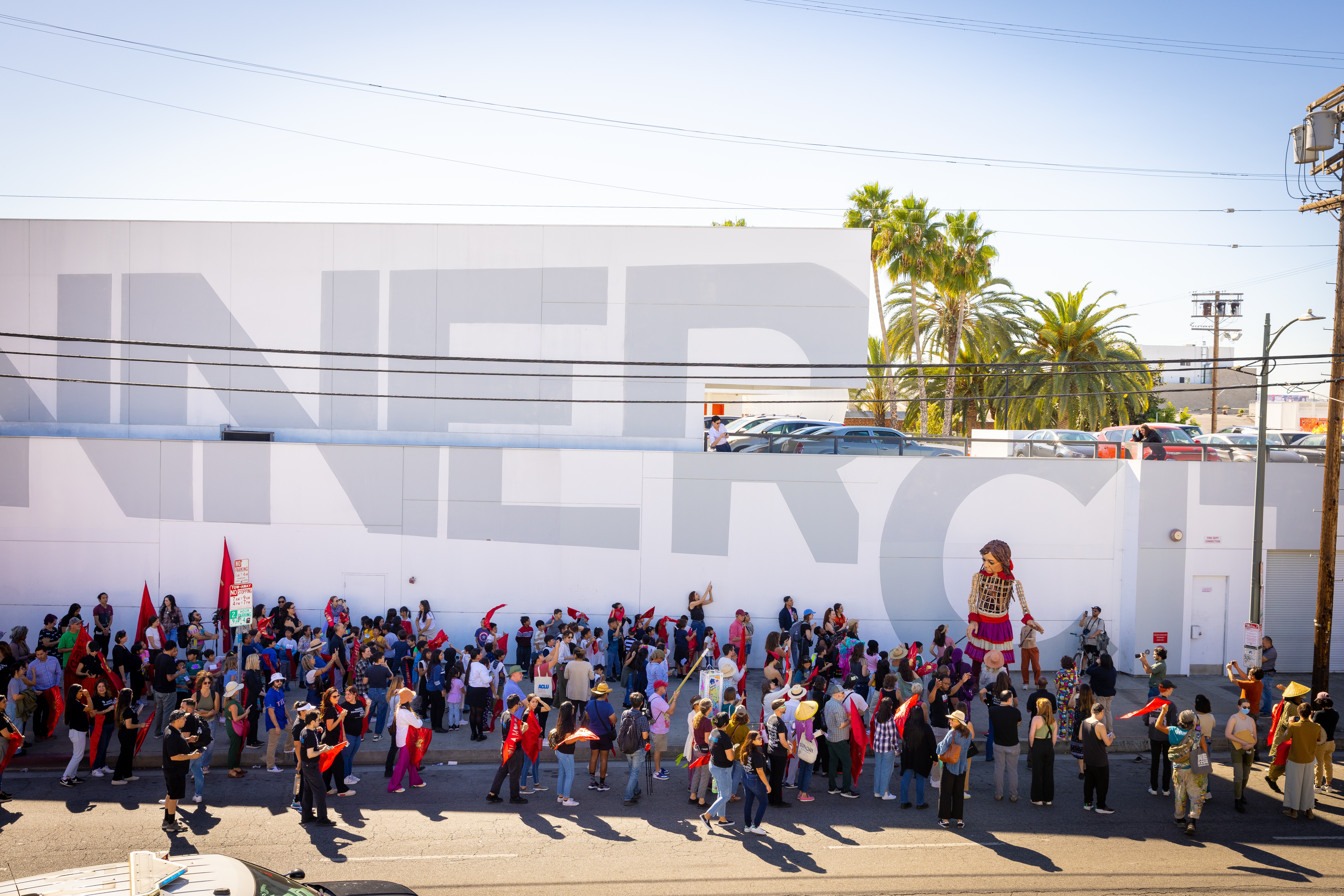 Crowd of people gathered outside a white wall with large gray letters, some holding umbrellas, in sunny weather.