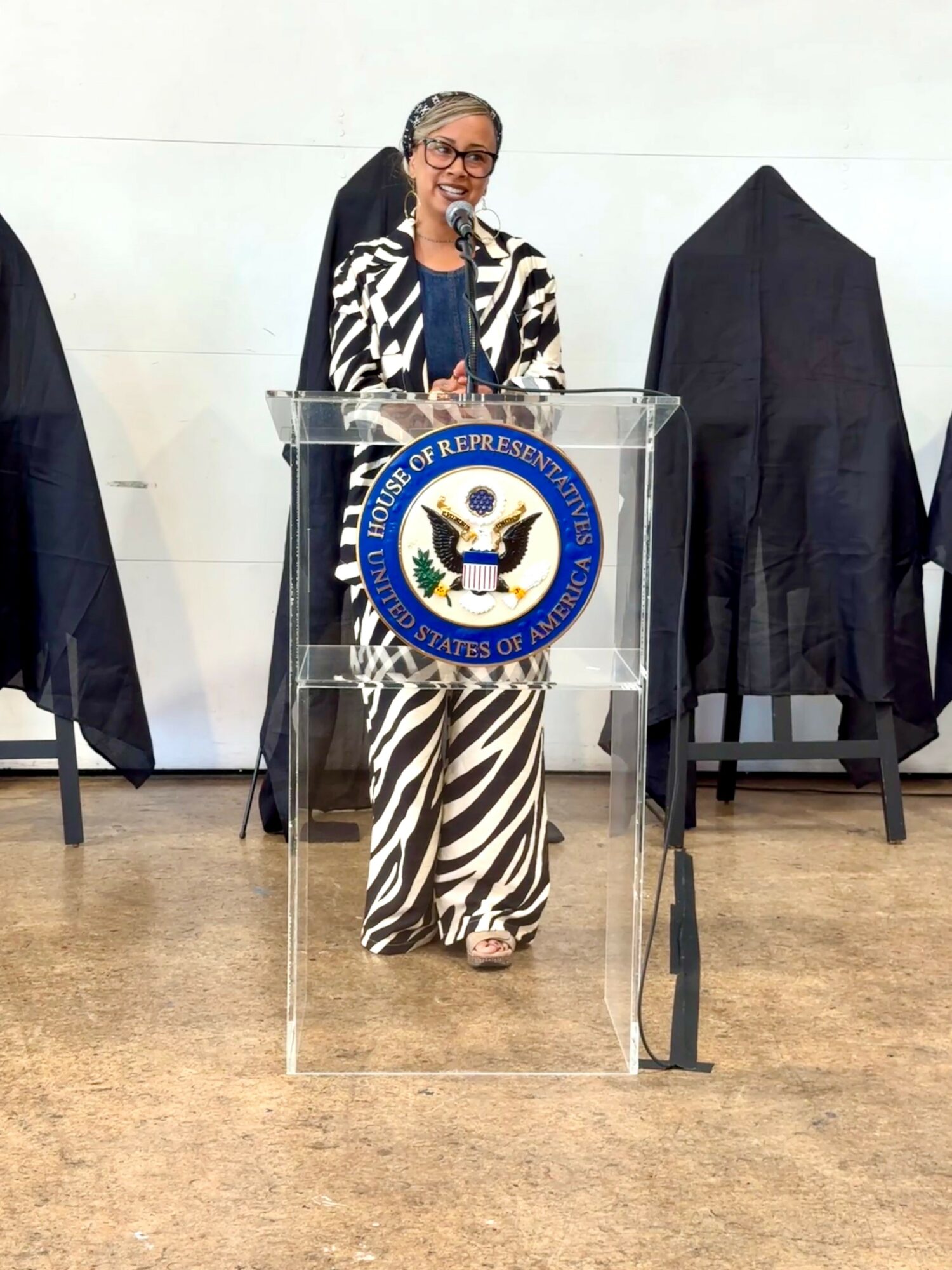 Woman standing at a podium with the seal of the U.S. House of Representatives, speaking into a microphone, wearing a zebra-patterned outfit.