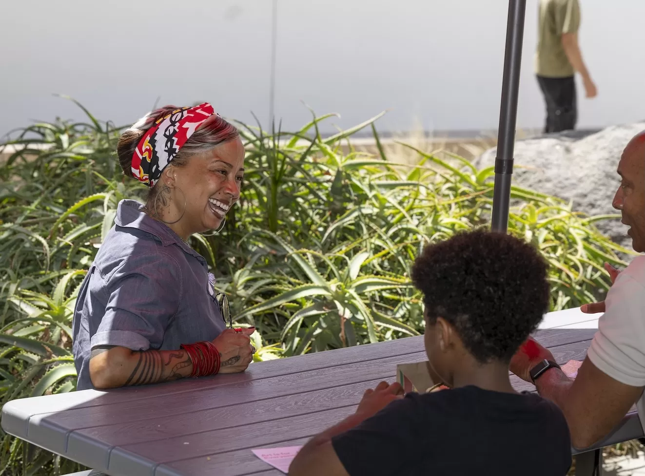 Three people sitting at a table outdoors, smiling and talking, with greenery and a person walking in the background.