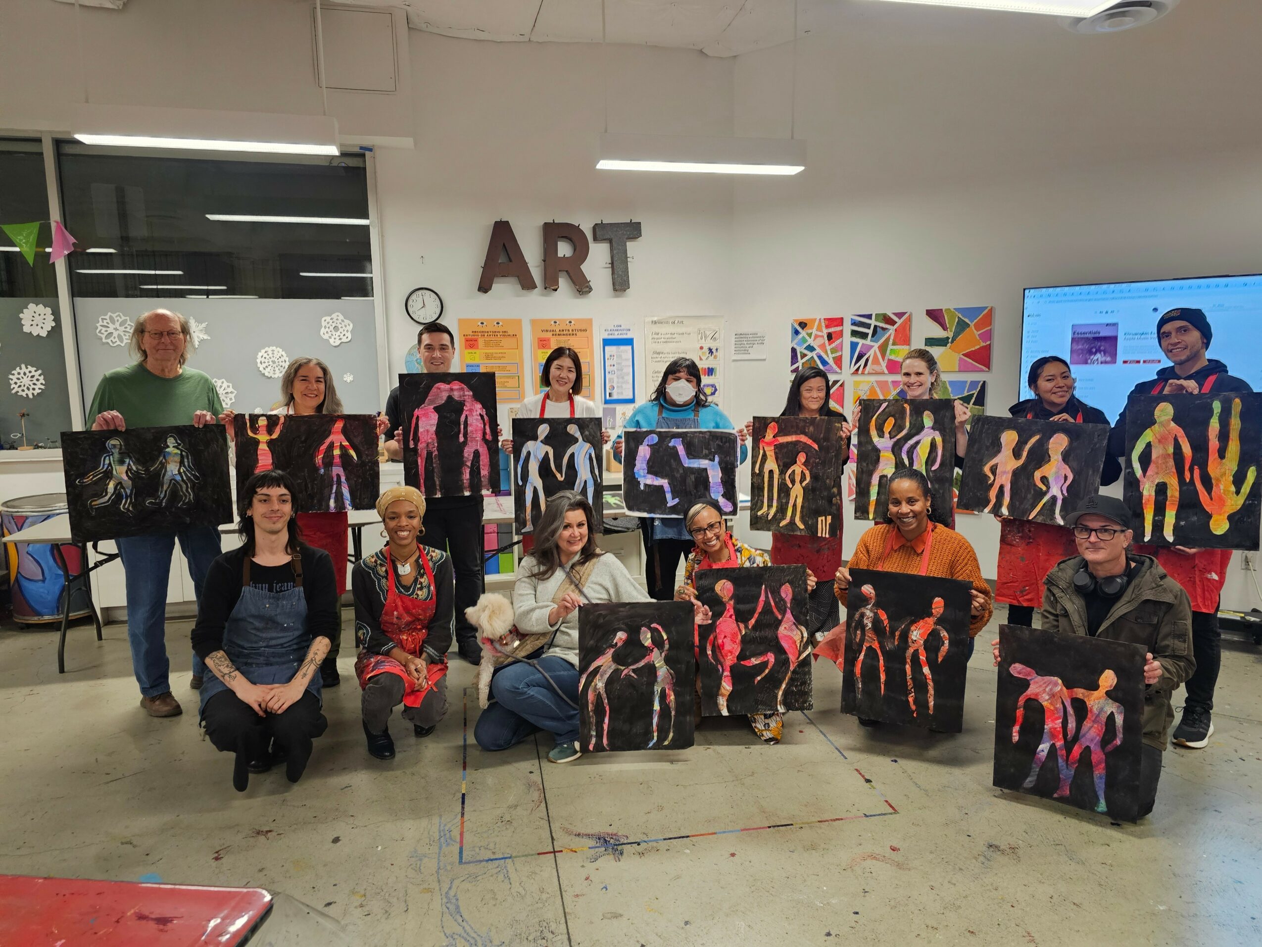 Group of people holding paintings with colorful abstract figures in an art studio, with a sign reading 'ART' on the wall.