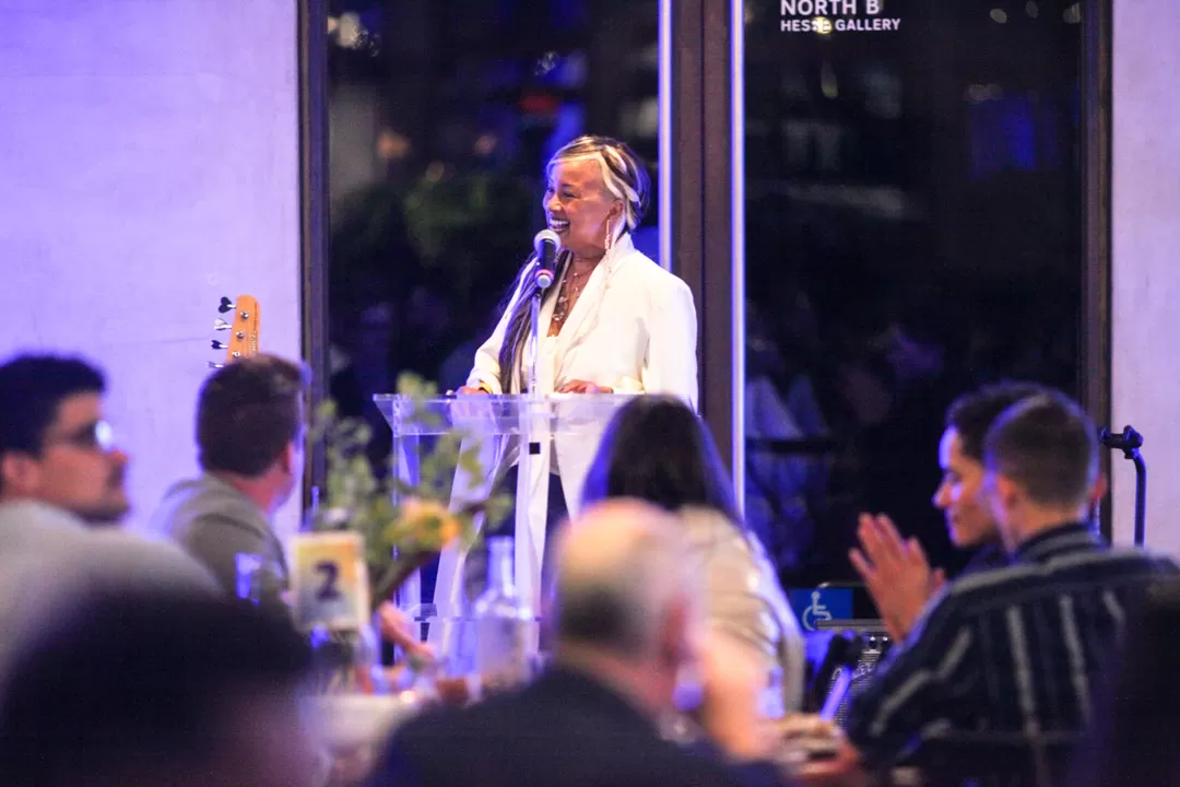 Woman speaking at microphone during event, audience seated at tables, some clapping, indoor setting with large window behind her.