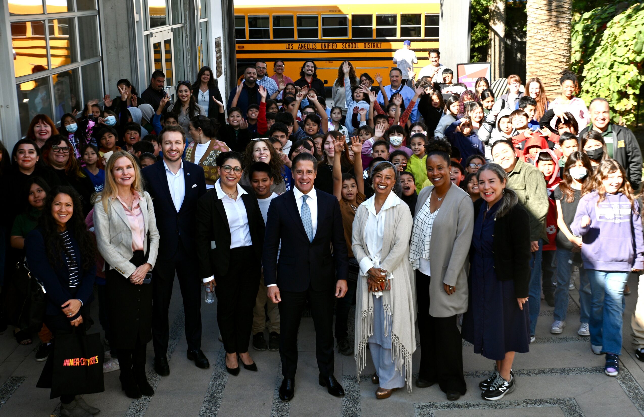 Group of diverse people standing outside a building, smiling, with a yellow school bus in the background.