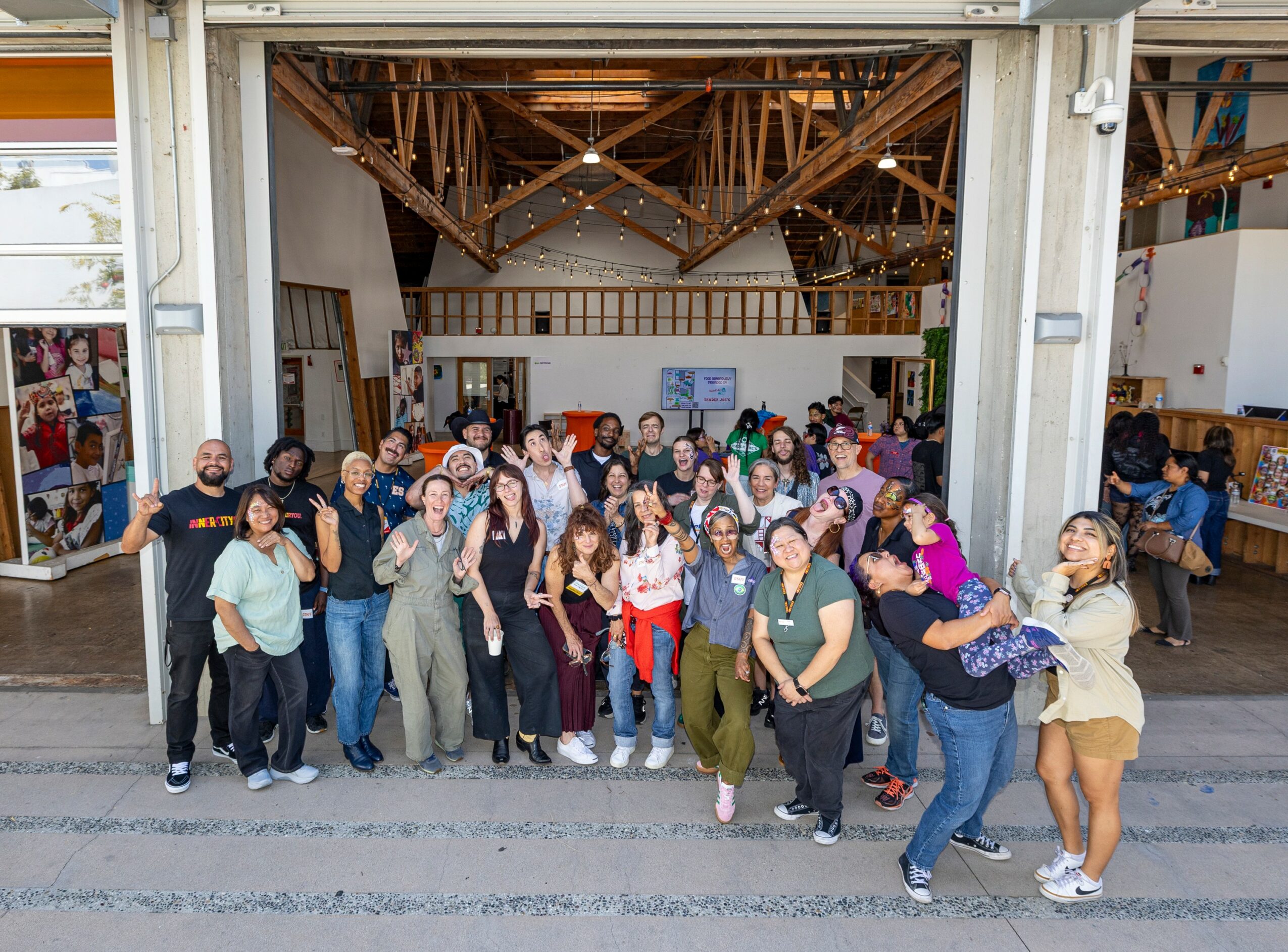 Group of people posing inside a large open building with wooden ceiling beams, some hugging, smiling, and waving.