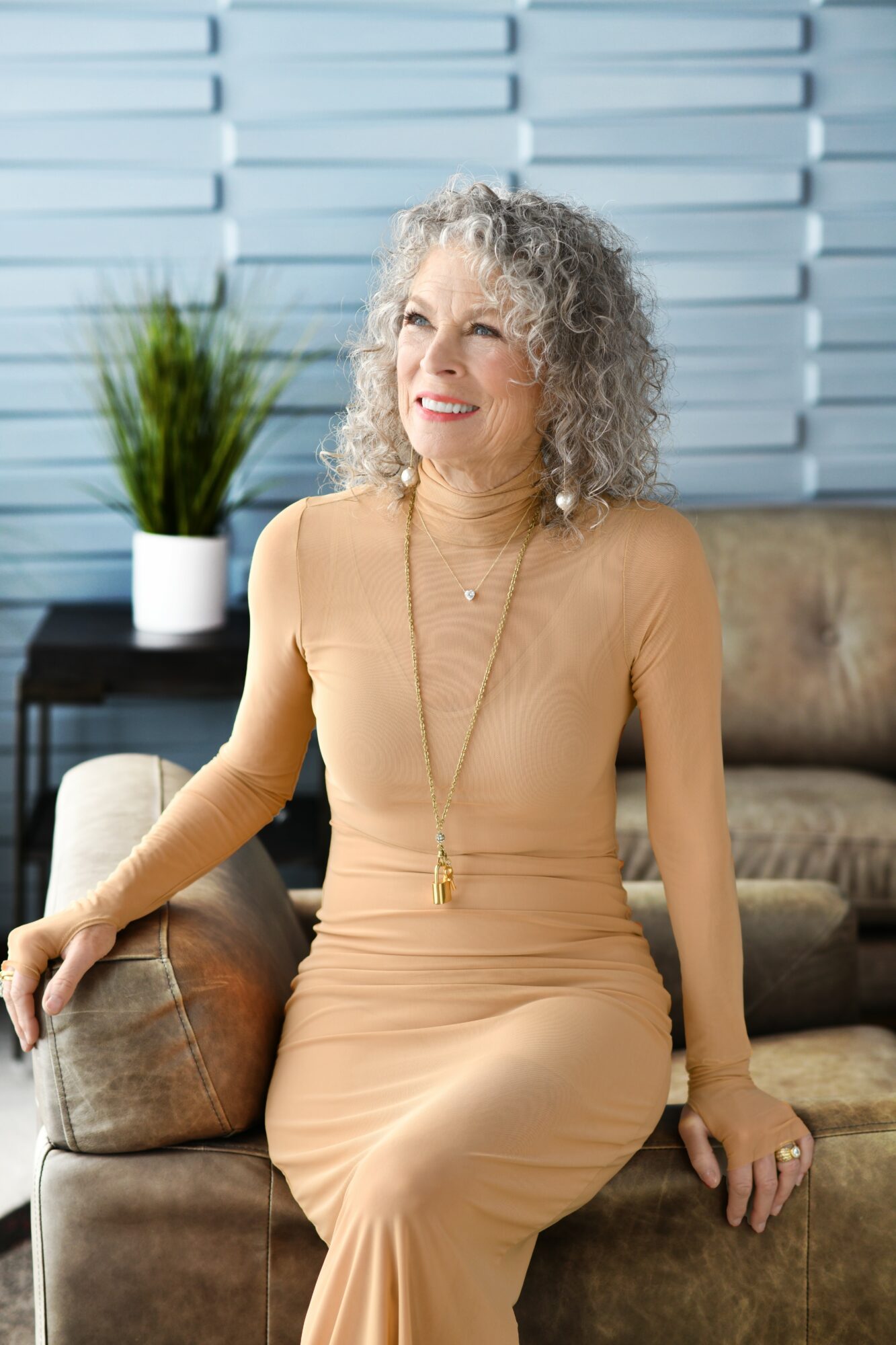 Older woman with curly gray hair sitting on a sofa, smiling, in a room with a plant and window blinds behind her.