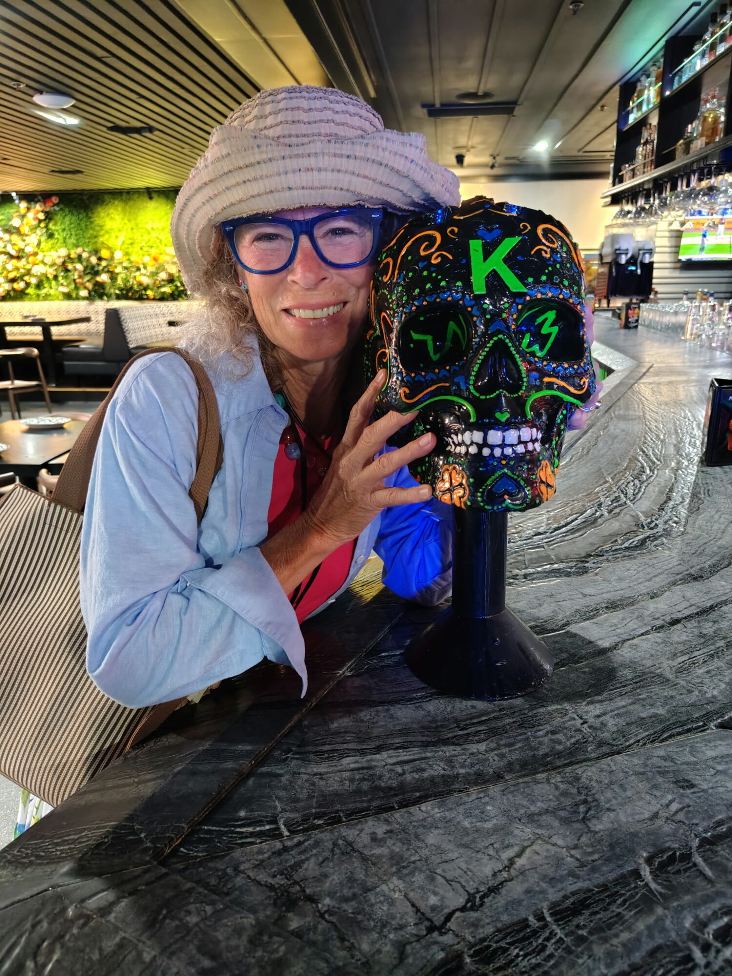 Woman with glasses and hat smiling next to colorful skull sculpture in a bar or restaurant setting.