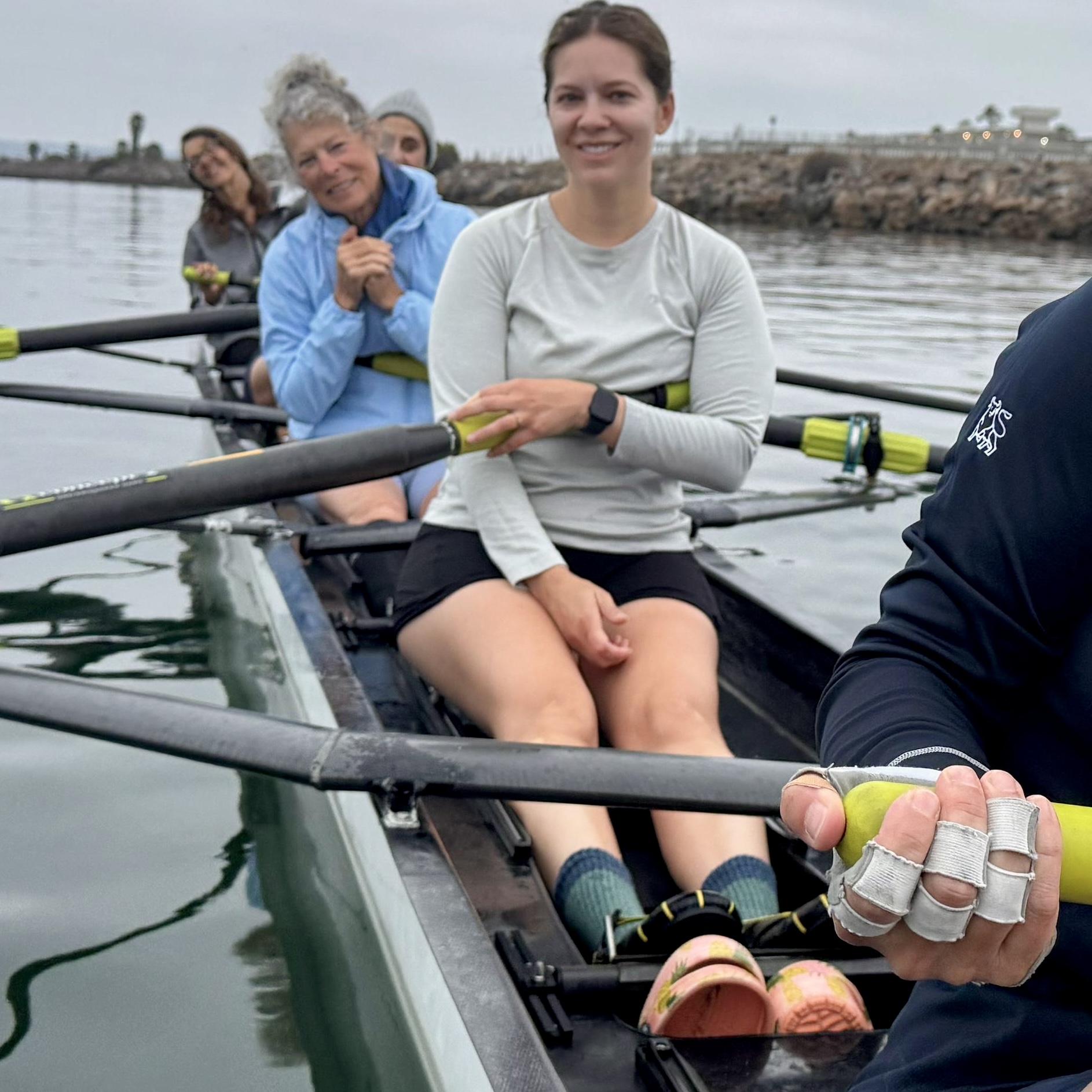 Group of women in a rowing boat on water, smiling and holding oars, with a dock in the background.