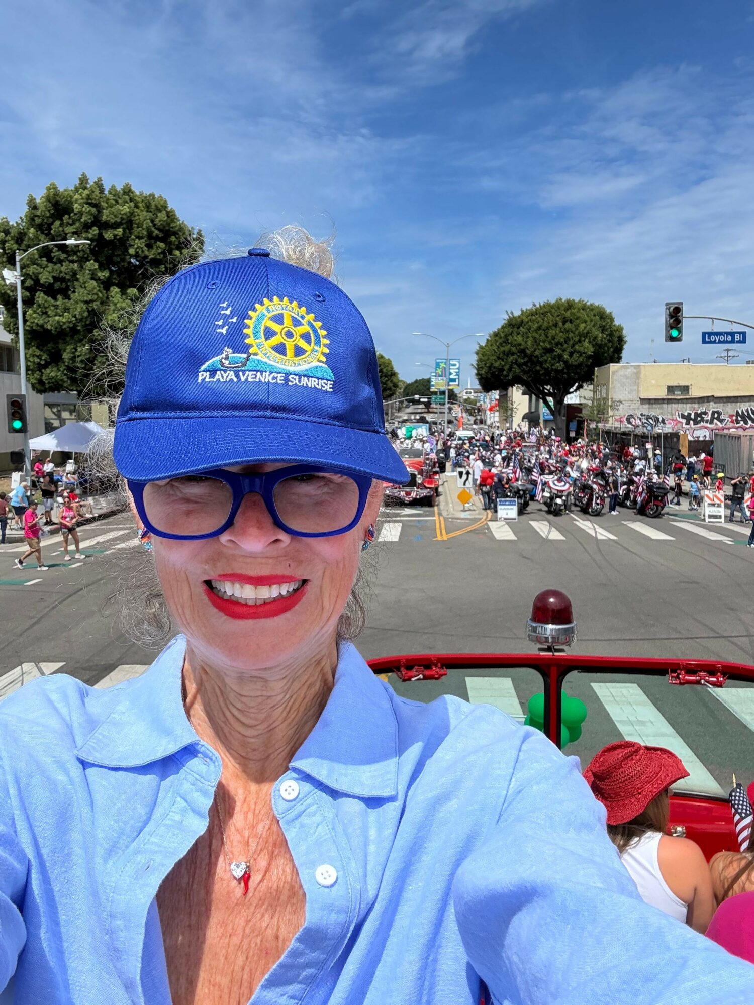 Woman wearing a blue cap and glasses, smiling outdoors with a crowd and tents in the background.