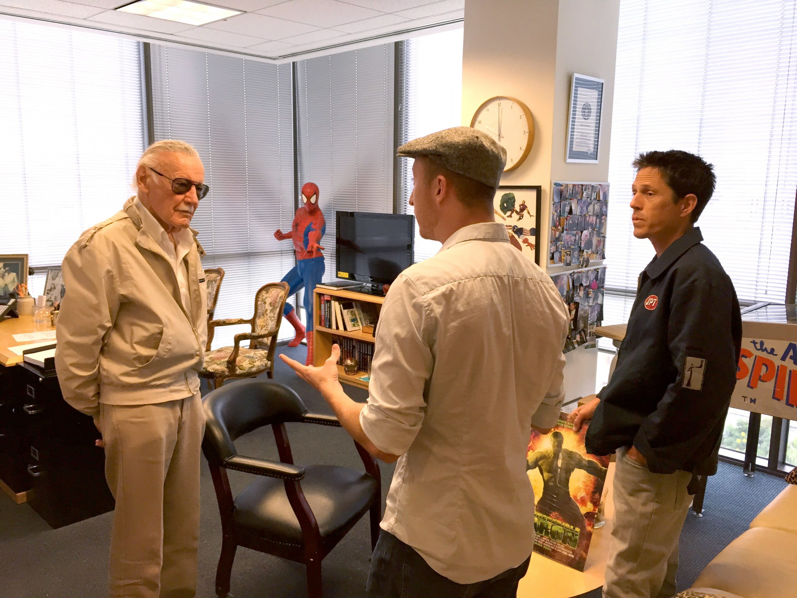 Three men are talking in an office with large windows, bookshelves, and a Spider-Man poster, engaged in conversation.