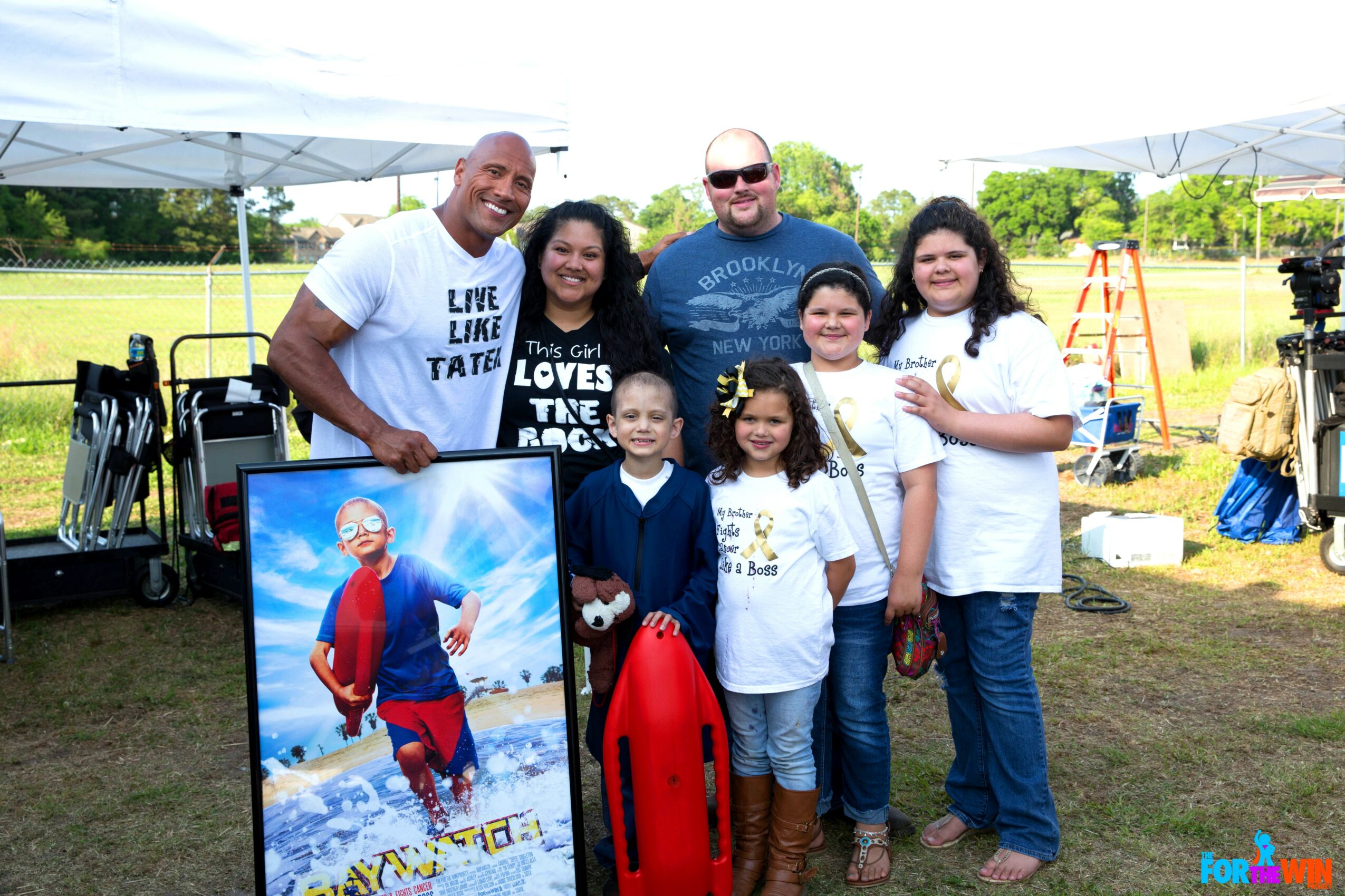 Group of six people outdoors, some children, standing near a large framed photo of a cartoon character, with tents and trees in background.