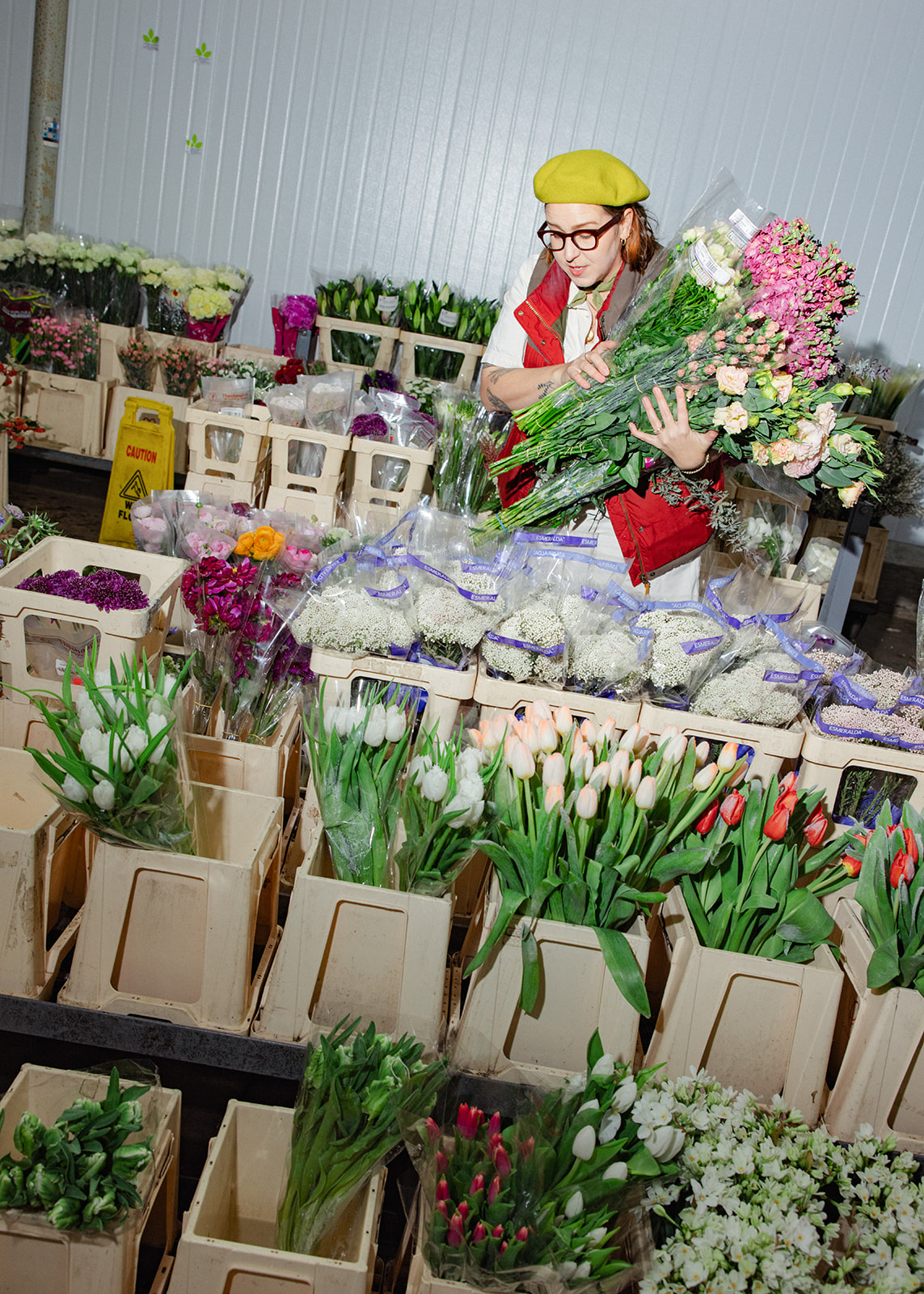 Woman in yellow hat and red apron arranging flowers in a flower shop, surrounded by various colorful flowers and plants.
