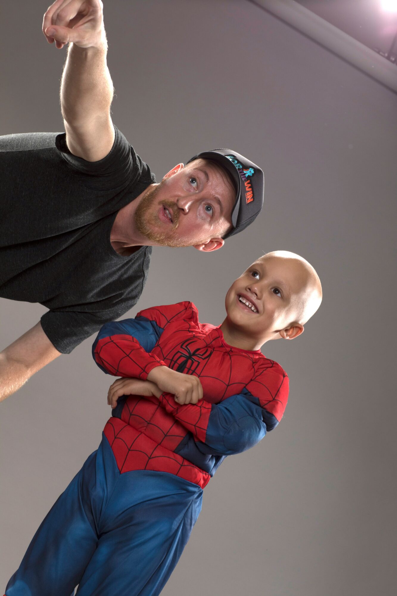 Man and child dressed as Spider-Man smiling and looking at the camera, indoors against a gray wall.