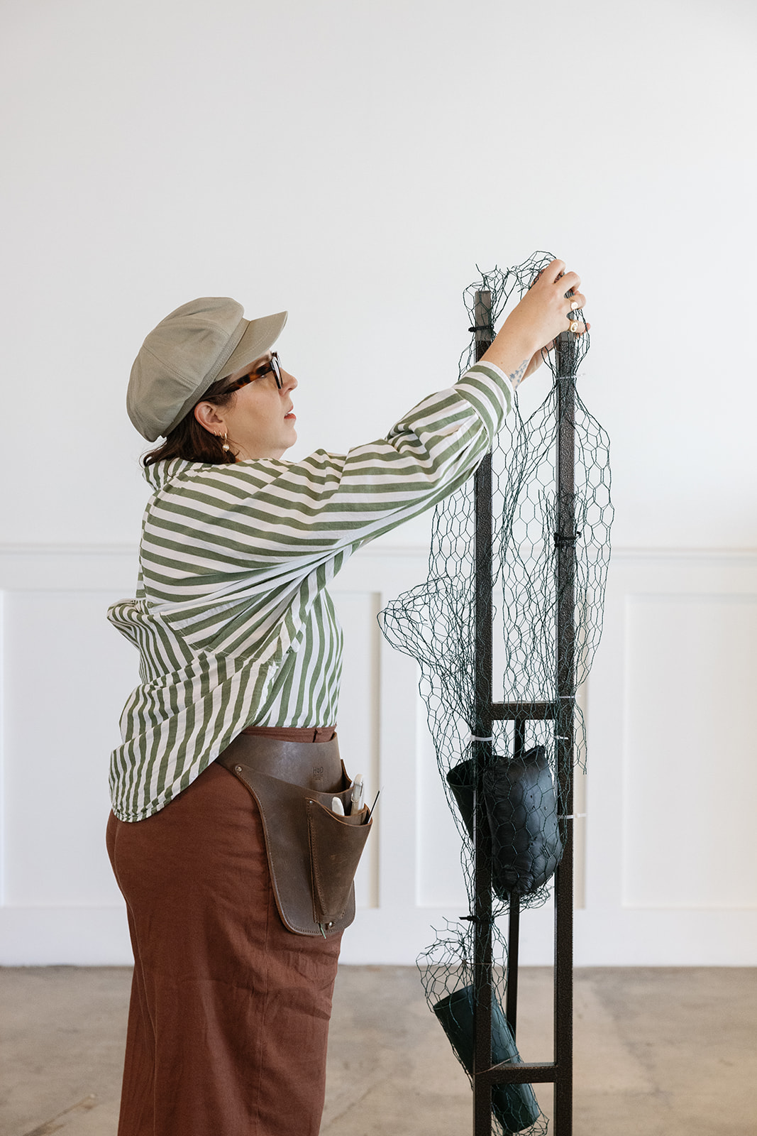 Person adjusting a tall wire mesh structure with a black bag hanging, standing indoors against a white wall.